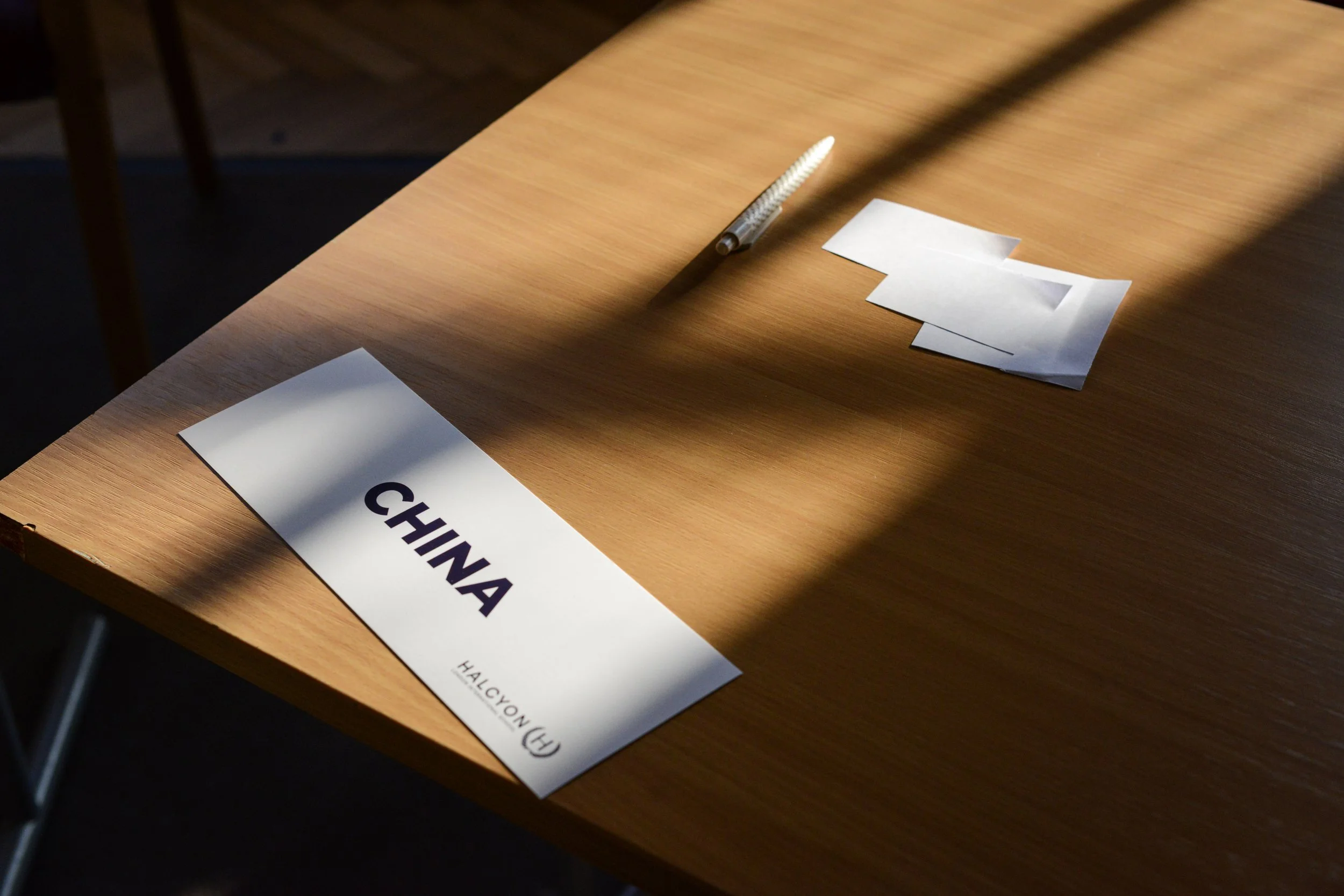 A wooden table with a white nameplate that says "CHINA" and a logo for Halcyon Fund, along with a white folded paper and a pencil on the table, with sunlight and shadows cast across the scene.