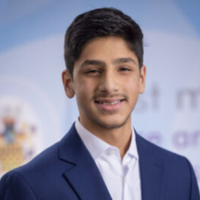 A young man in a navy blue suit and white shirt with a slight smile, standing outdoors with a blurred background.