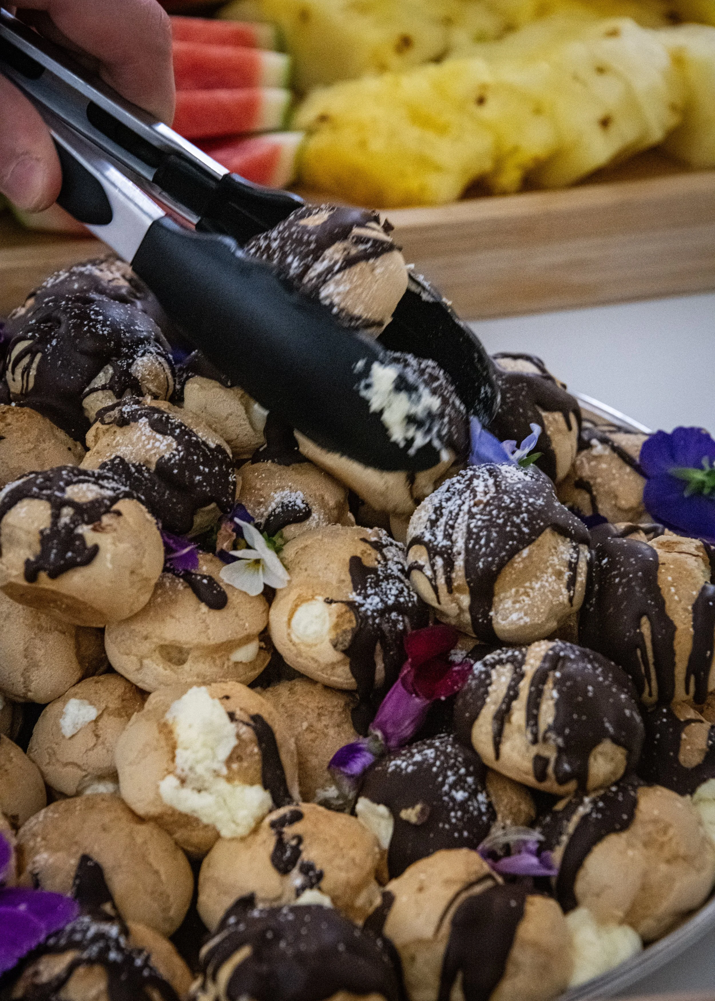 A tray of cream puffs drizzled with chocolate sauce and topped with edible flowers.
