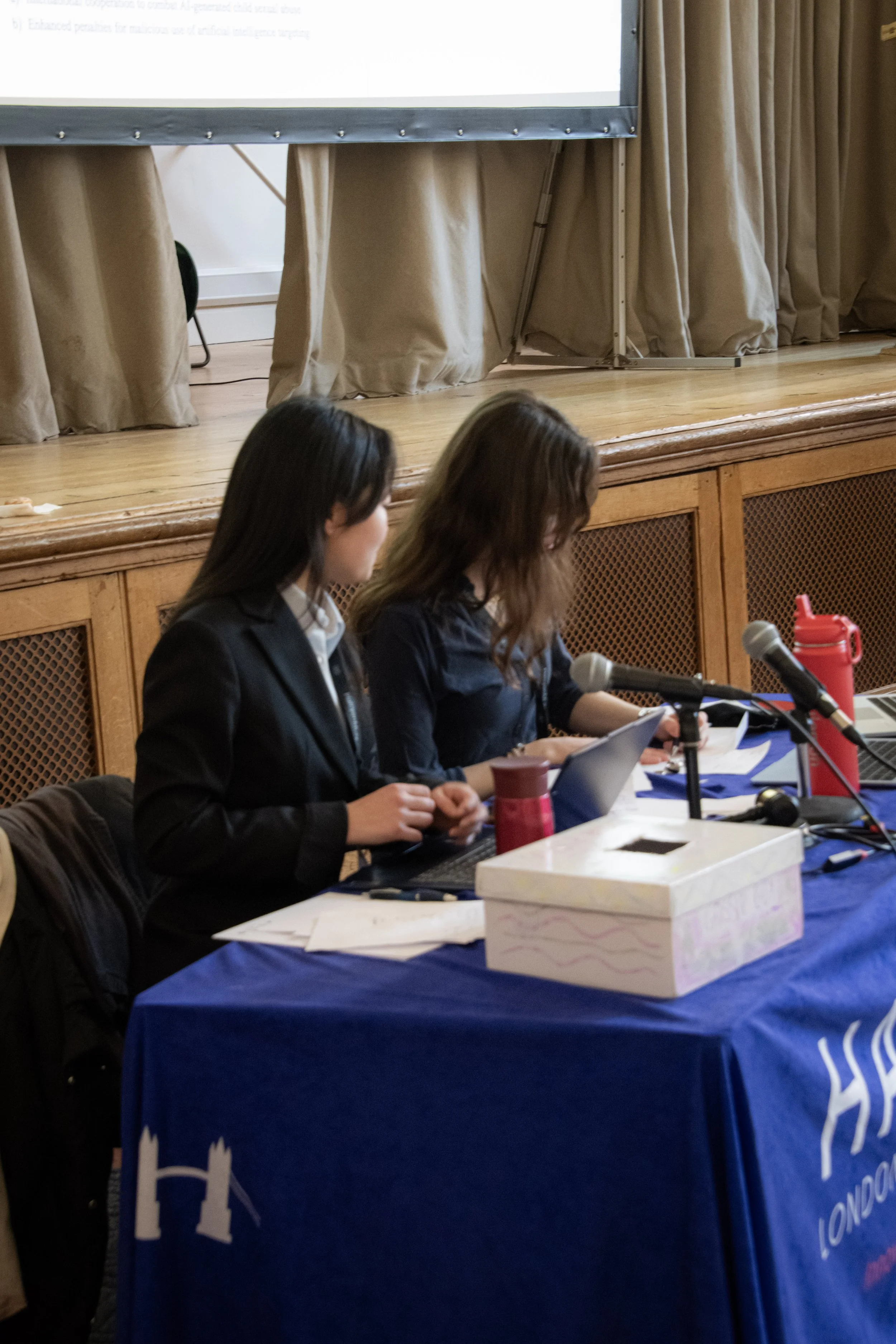 Two women are sitting at a table with microphones, in front of a large screen, possibly during a conference or panel discussion.