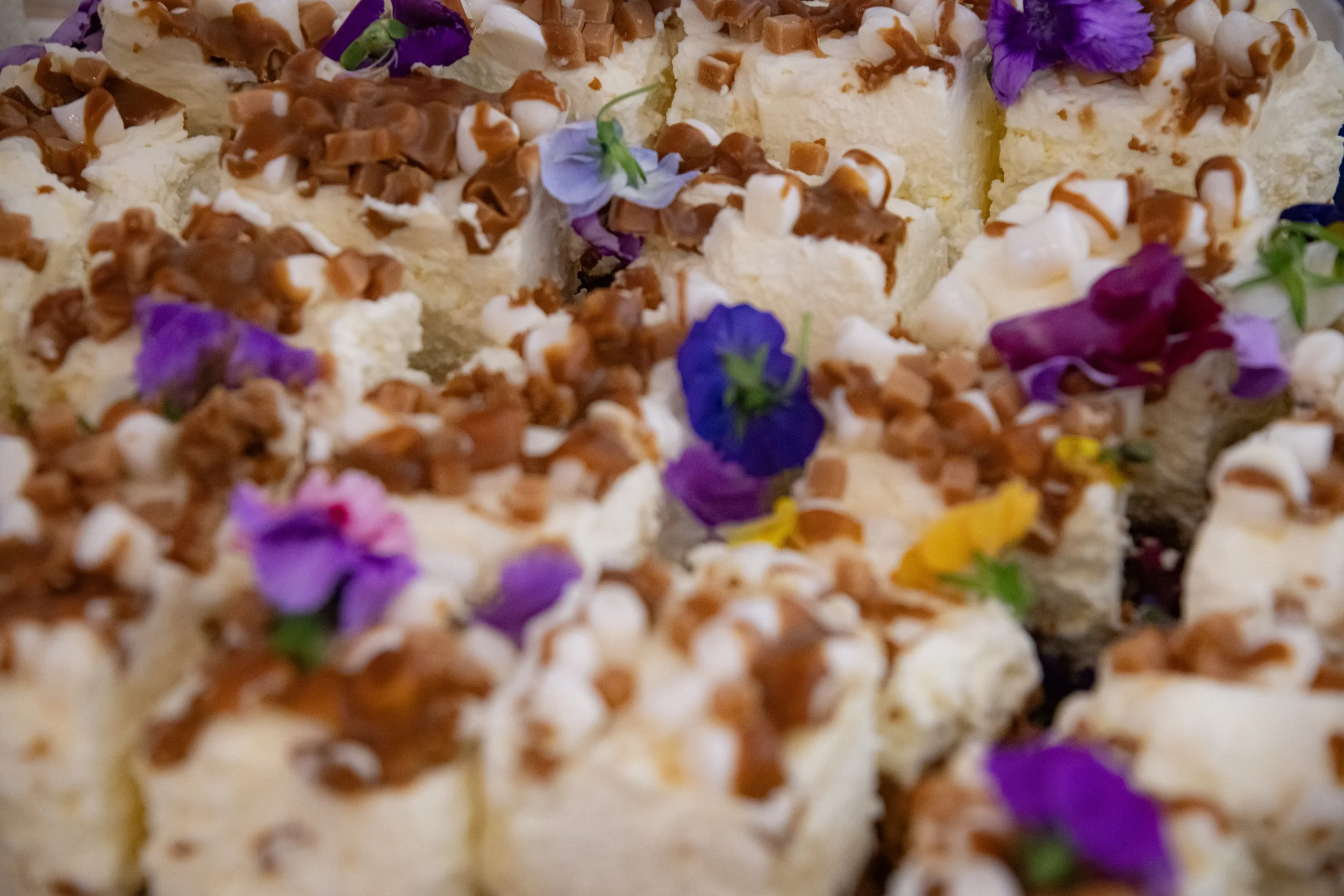 Close-up of white cake squares topped with chocolate drizzle and colorful edible flowers.