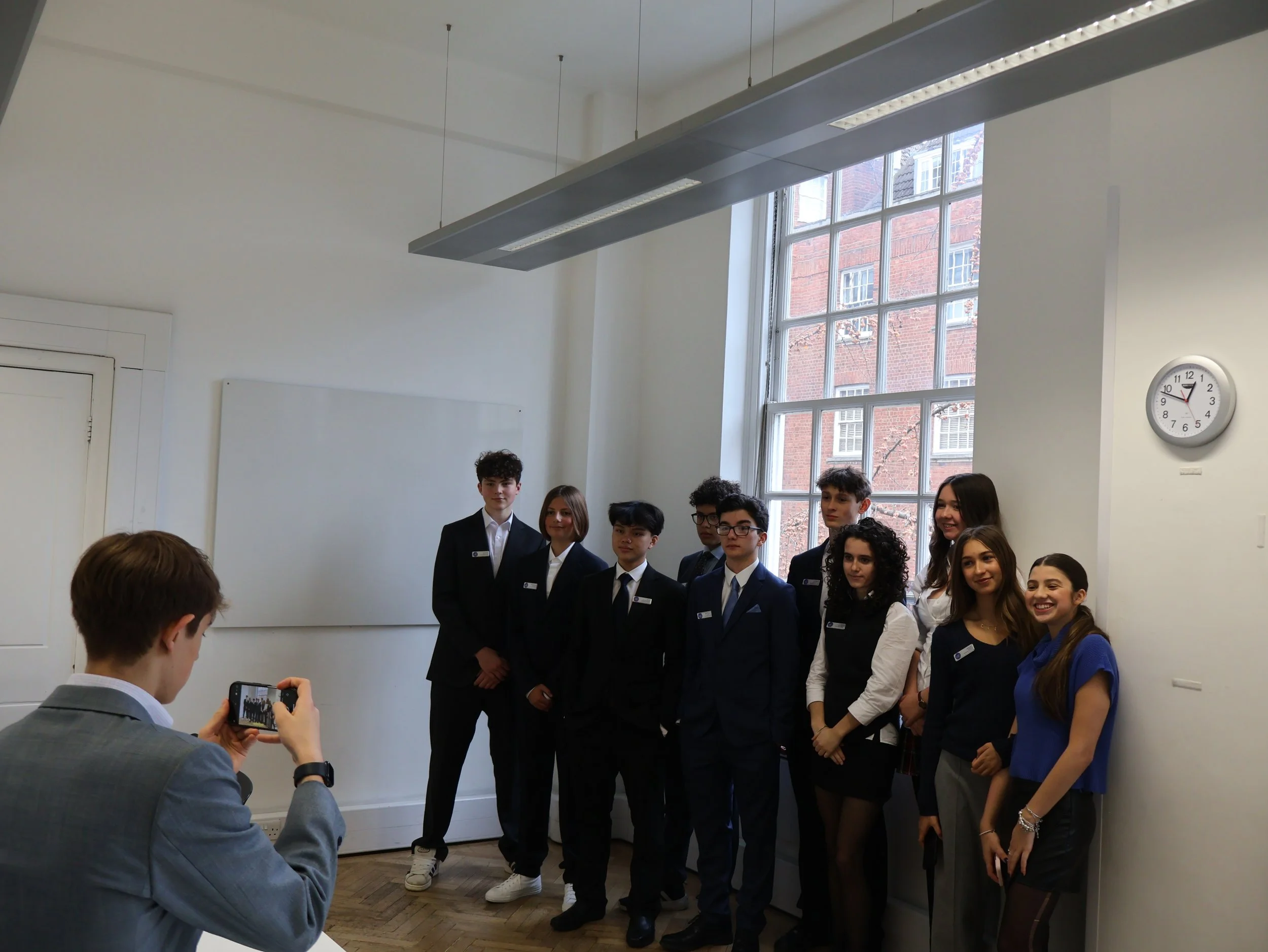 Group of students in formal attire posing for a photo in a classroom, with a teacher or organizer taking their picture. The room has large windows, a whiteboard, and a wall clock showing 12:50.