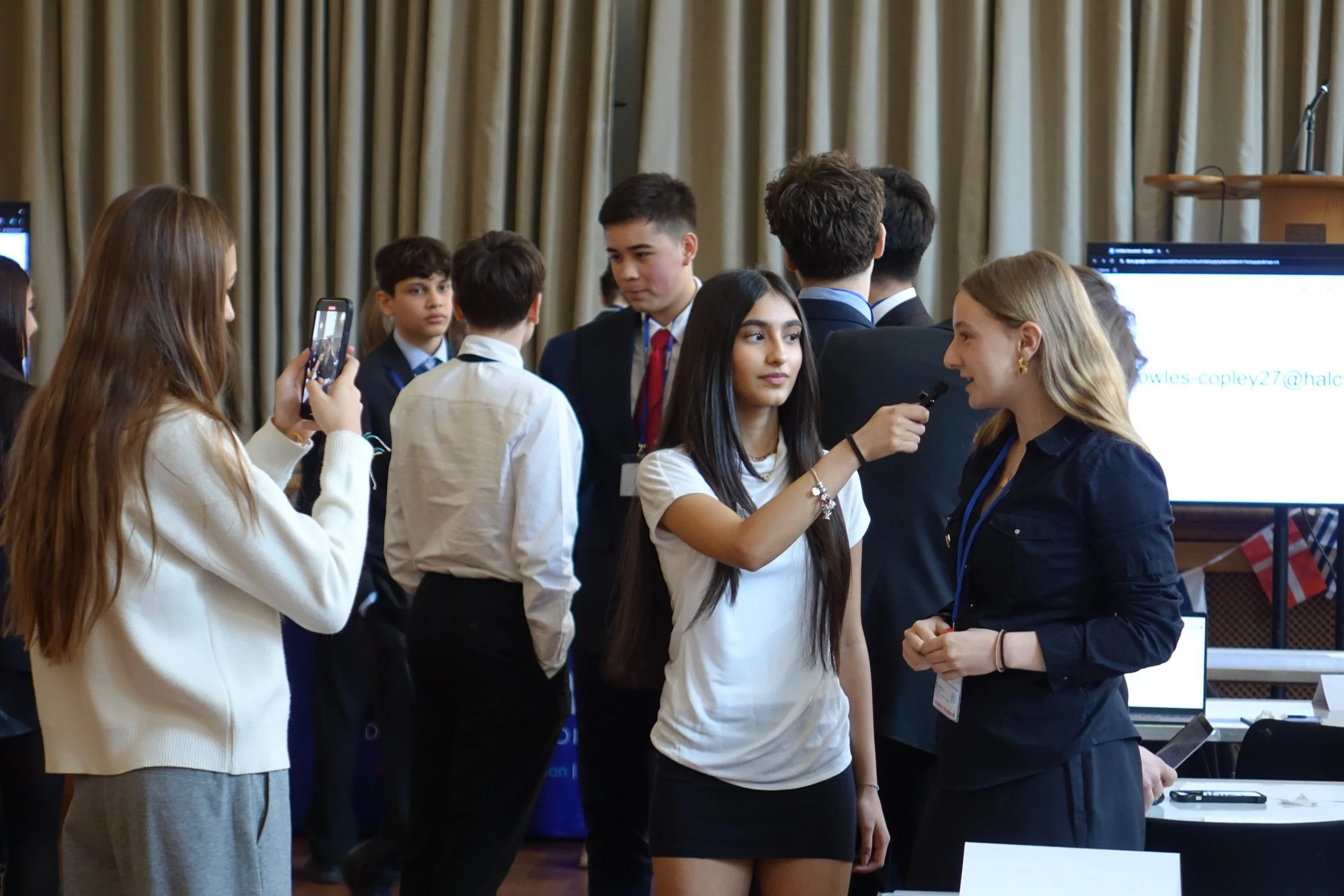 Young woman being interviewed at a conference or event with other young people and presentations in the background.