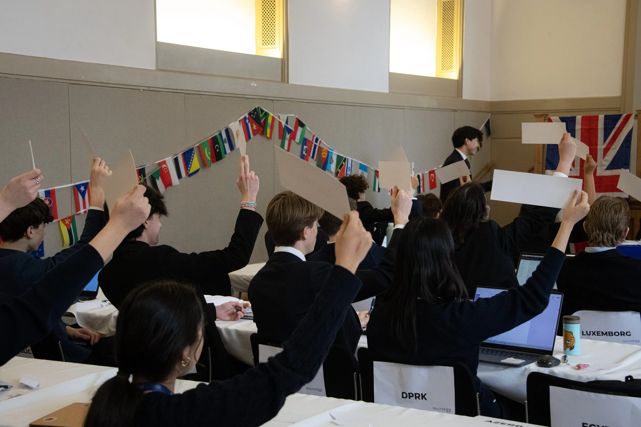 Young people participating in a Model United Nations conference, raising their hands in a voting session with national flags and a British flag in the background.