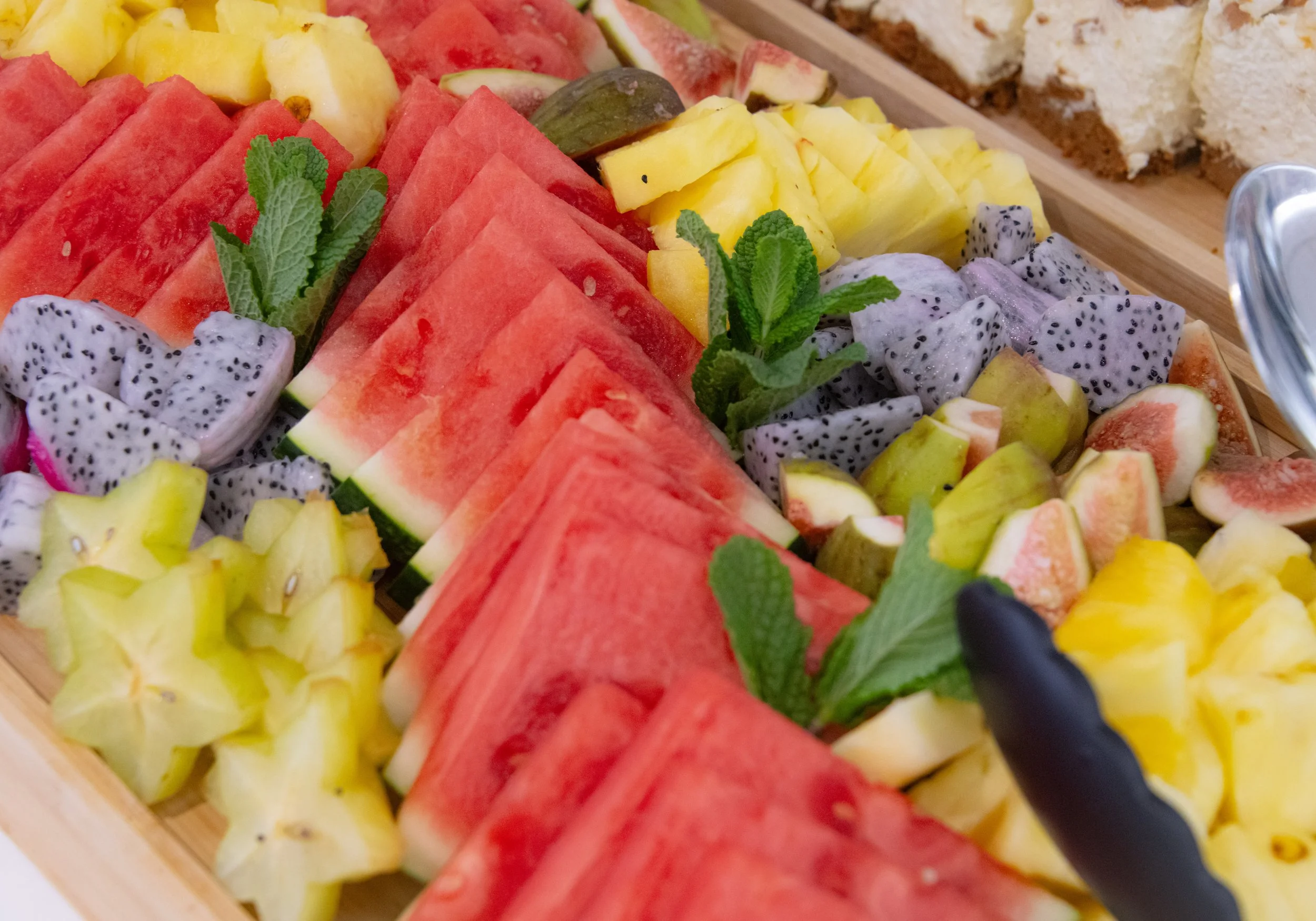 A tray of assorted fresh sliced fruits including watermelon, pineapple, dragon fruit, fig, and honeydew, garnished with mint leaves.