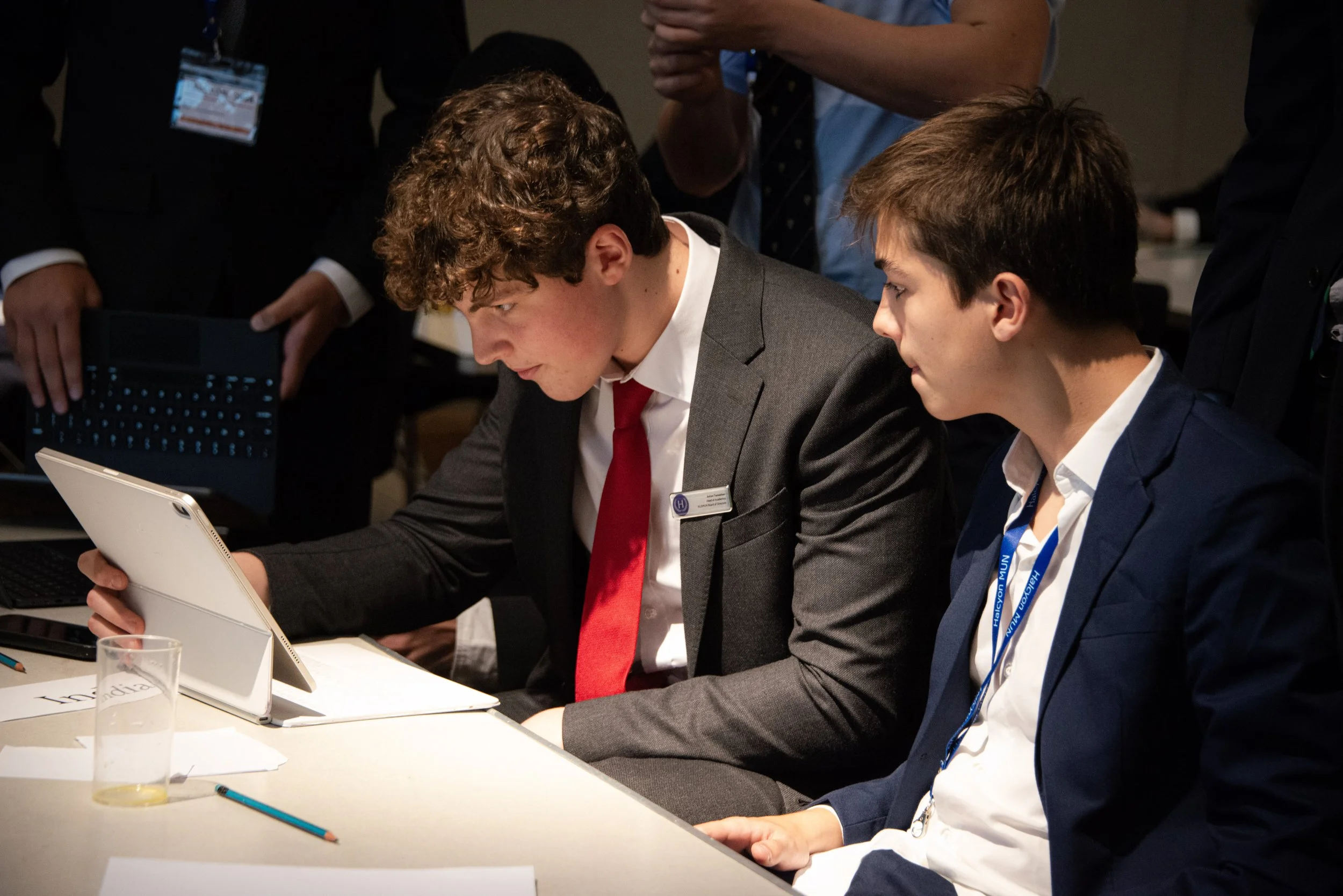 Two young men seated at a table working on a tablet, with other people standing around them in a professional setting.