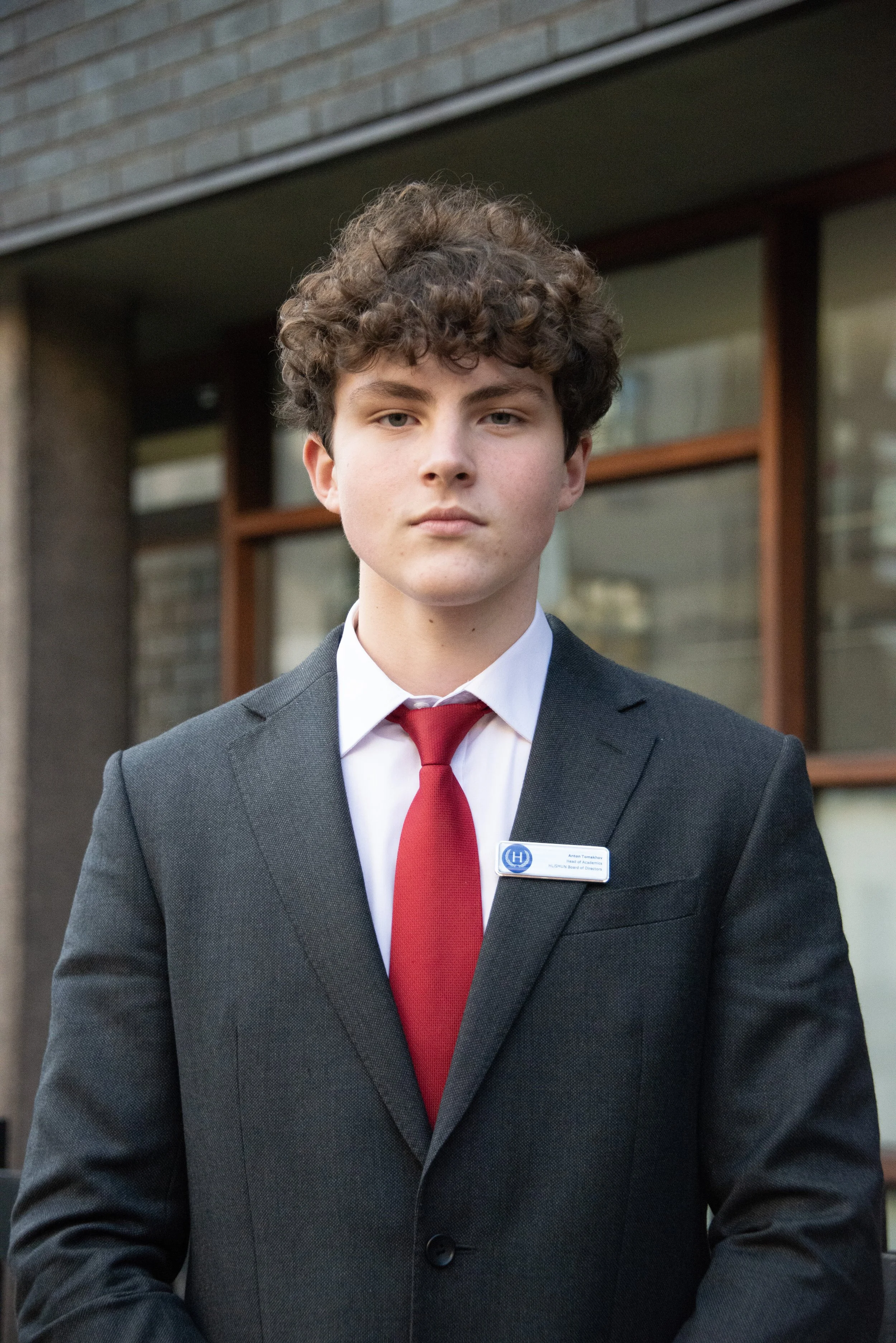 A young man in a black suit with a red tie and white shirt standing outside a building with large windows and brick walls.