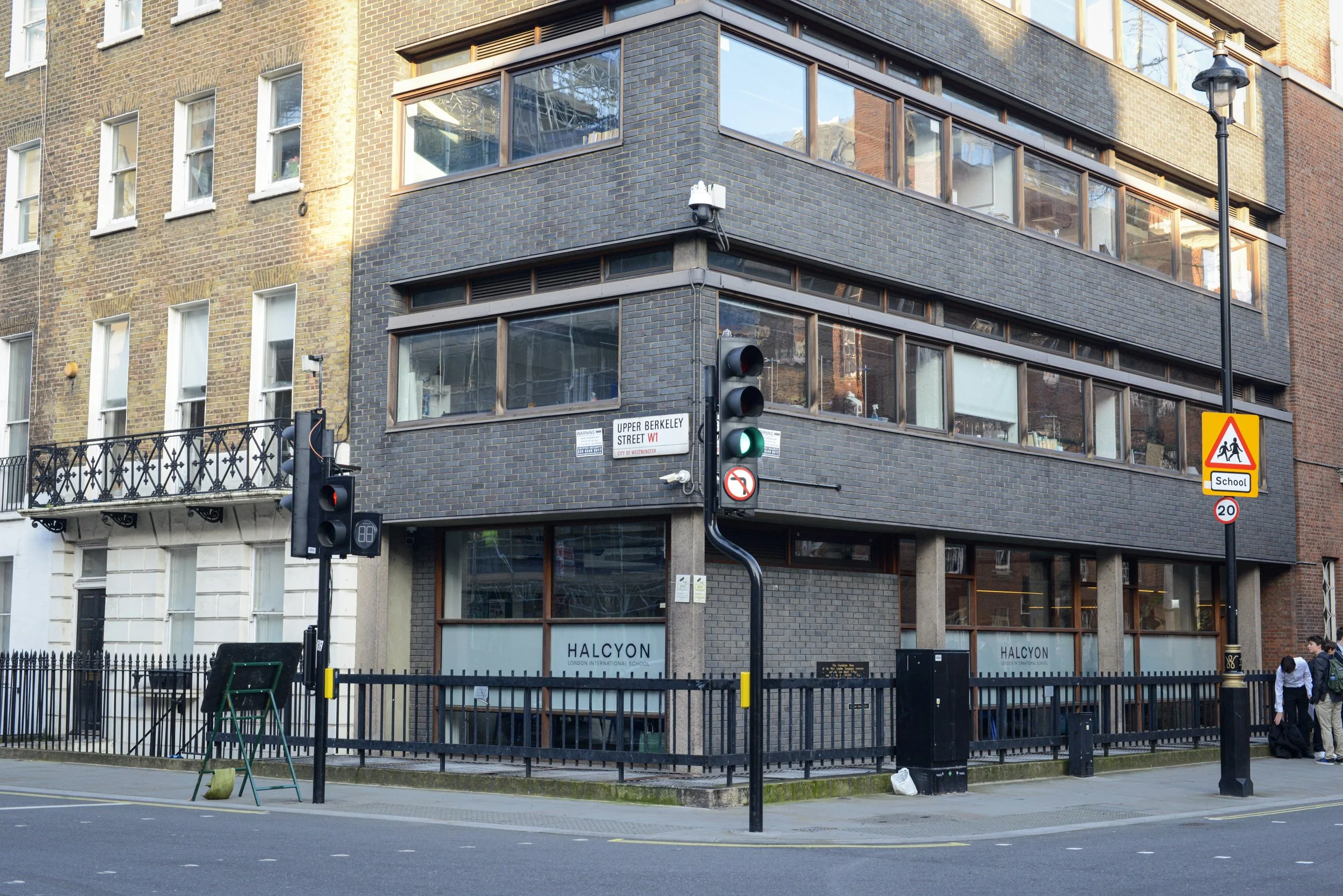 Street corner with traffic lights, school zone sign, and a building with large windows and a black brick facade. There are pedestrians on the sidewalk and a fence around the building.