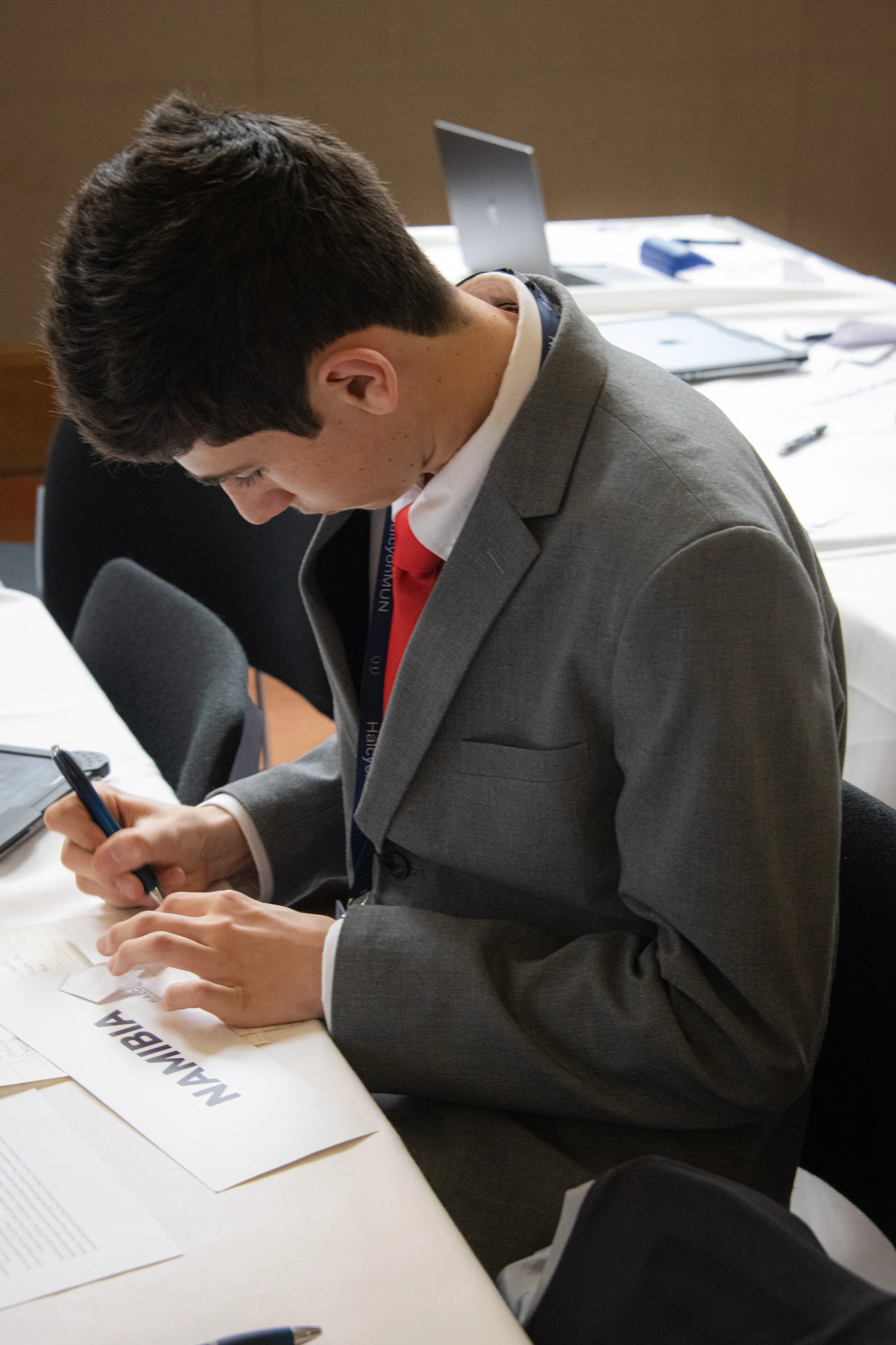 A young man in a grey suit and red tie sitting at a conference table, writing on a paper labeled "MAAMIBA."