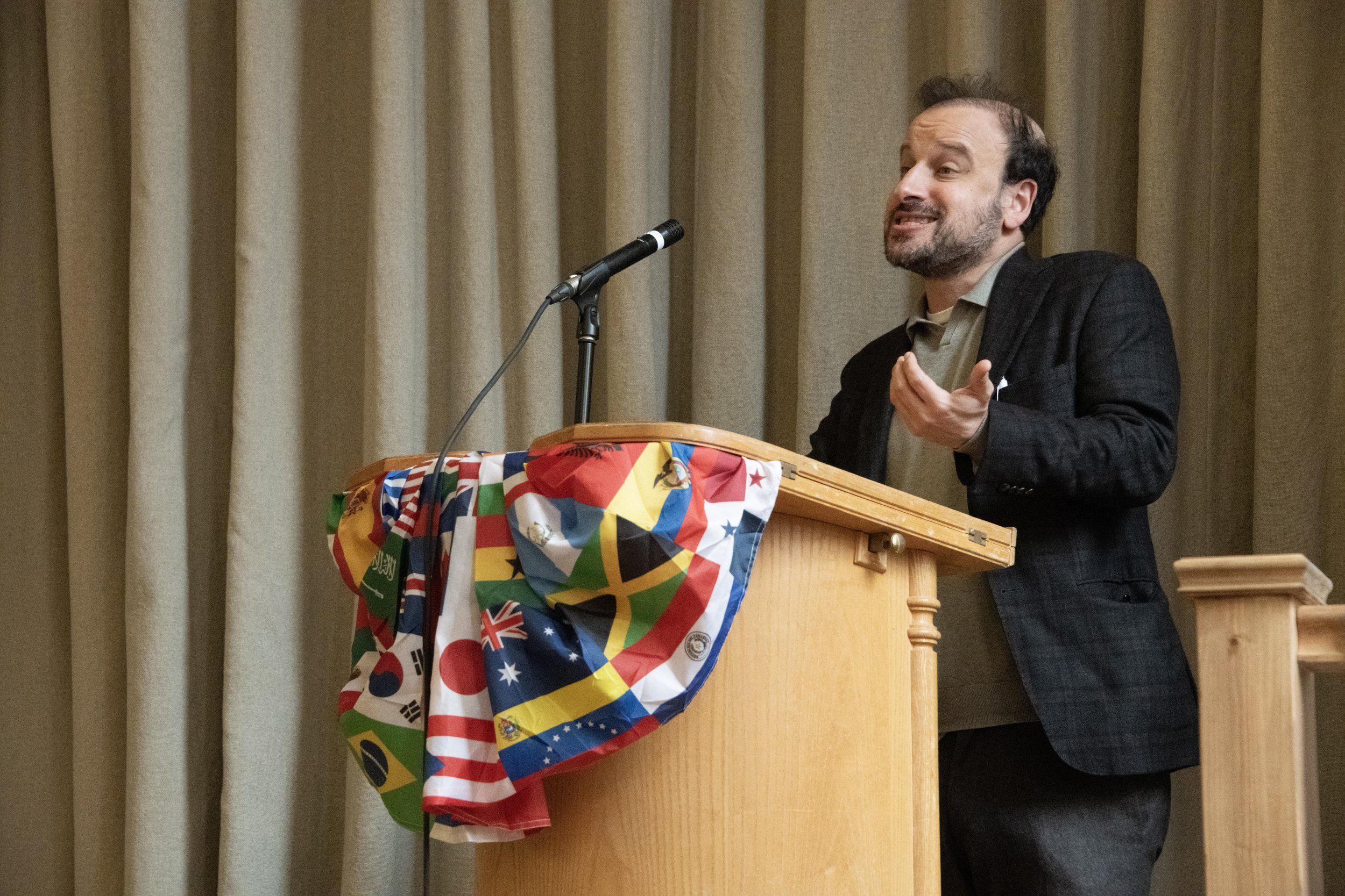A man with a beard and short hair wearing a dark blazer speaking at a podium draped with multicolored flags, in front of beige curtains.