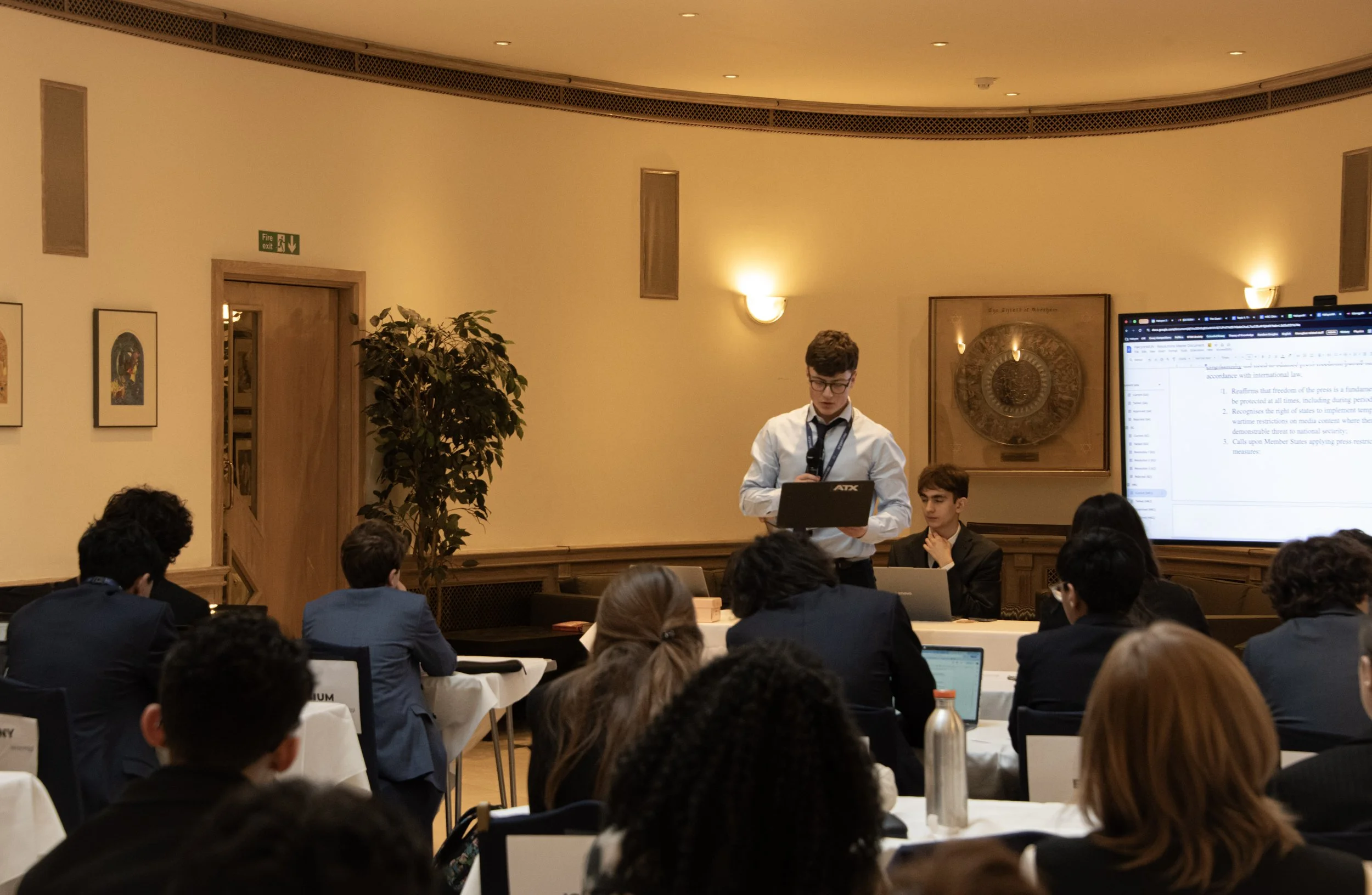 Young man in a white shirt presenting at a conference with a laptop, a large screen displaying text, and an audience seated at tables in a well-lit room.