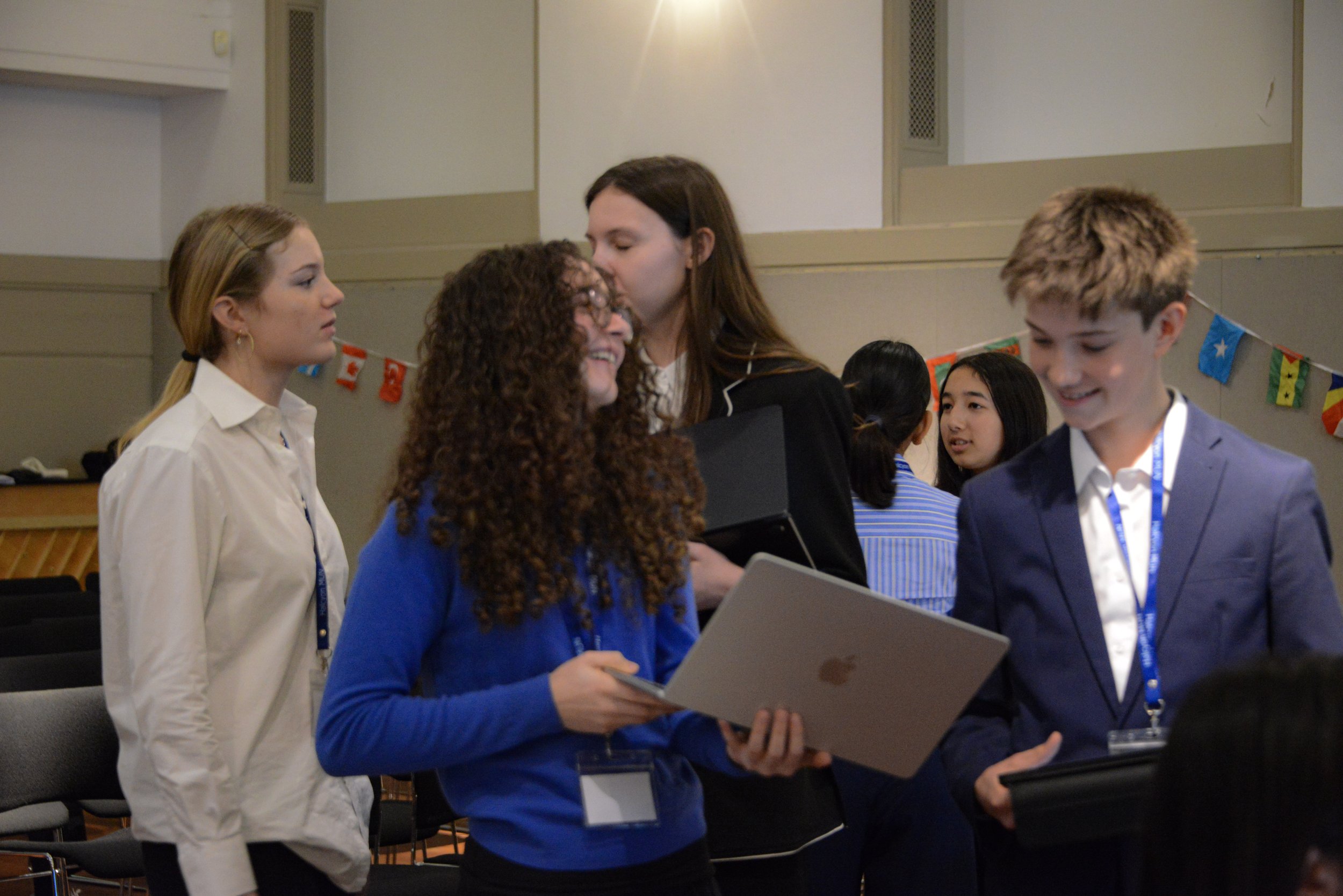 Group of young professionals at a conference or meeting, engaging in conversation and smiling, with international flags decoration in the background.