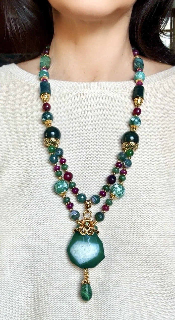 Close-up of a woman wearing a multicolored beaded necklace with large green and blue pendant on a beige top.