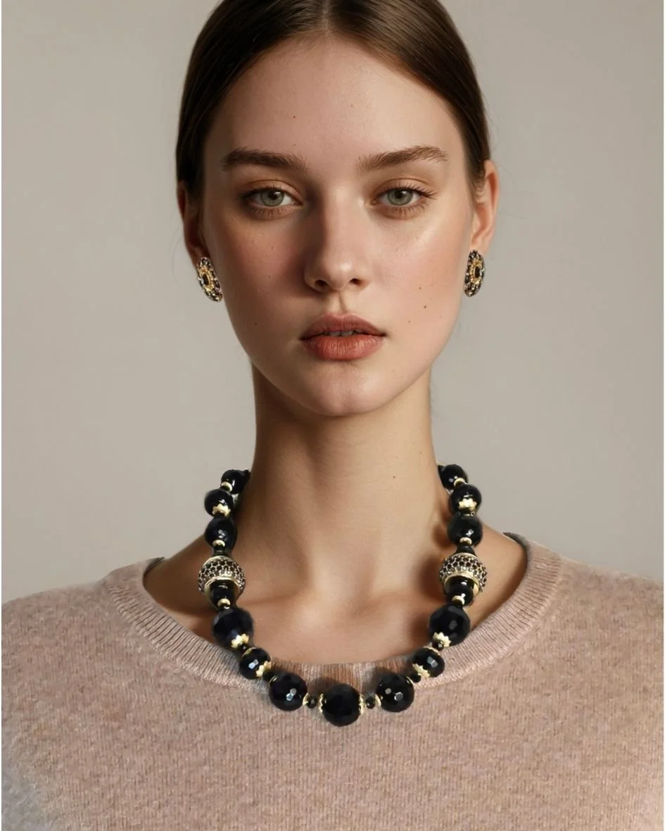 A young woman with fair skin and short brown hair wears a beige sweater, gold and black earrings, and a matching black bead necklace with gold accents, looking directly at the camera against a plain background.