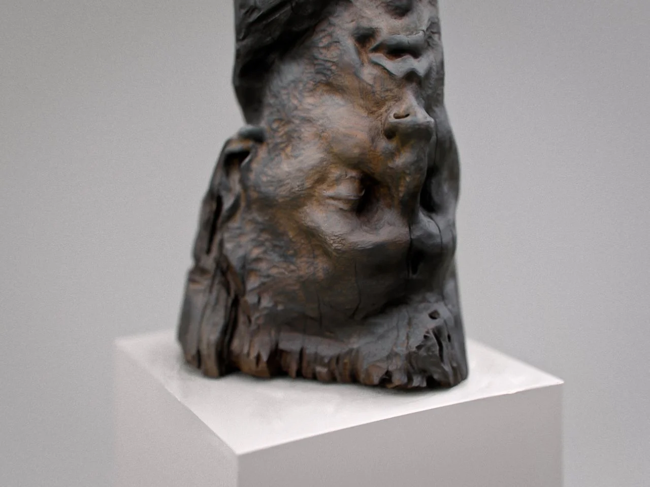 Close-up of a dark wood sculpture of a man's face upside down on a white pedestal, with detailed facial features and textured surface.
