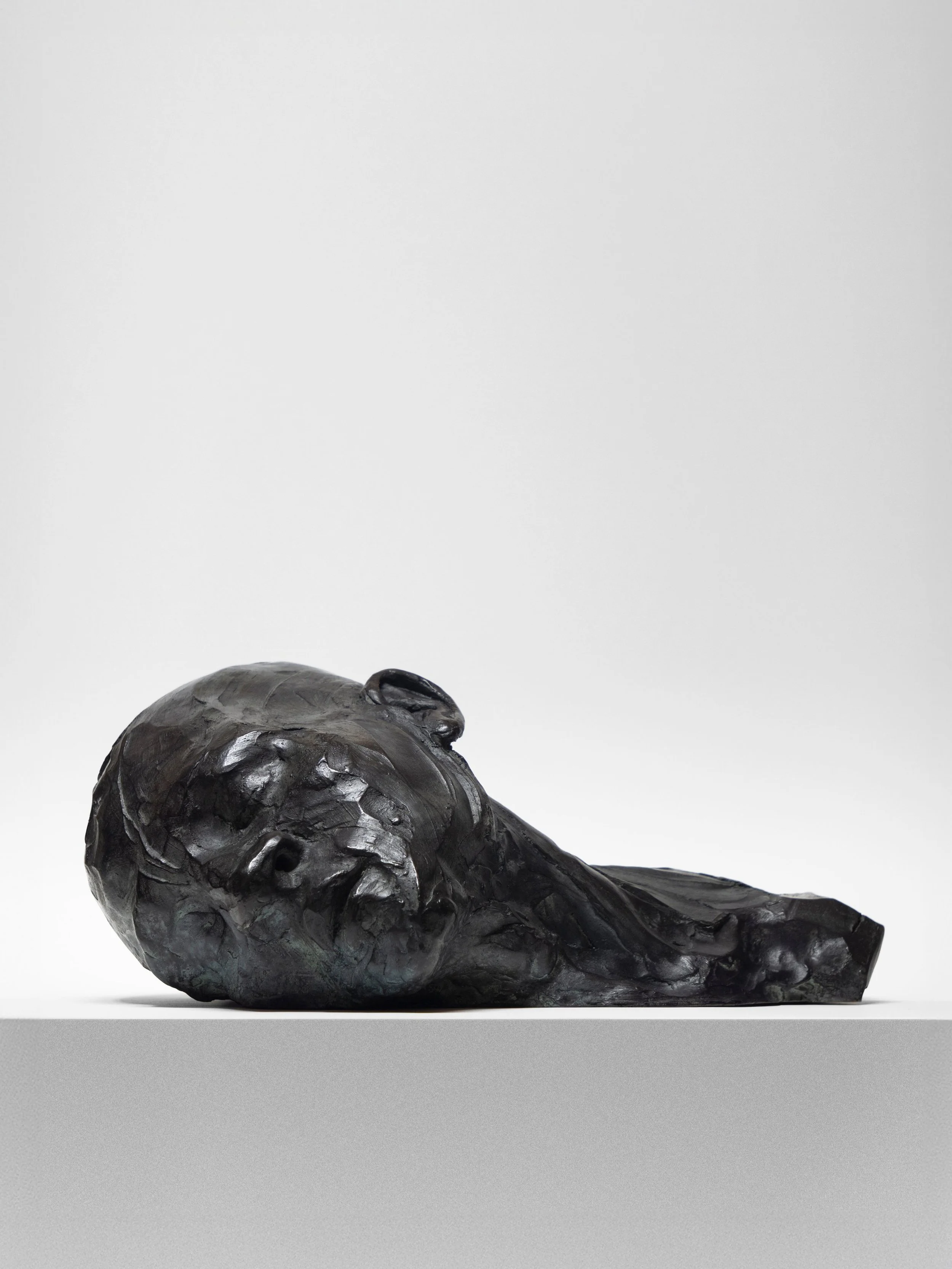 Dark bronze sculpture of a human head lying on its side on a white surface against a plain white background.