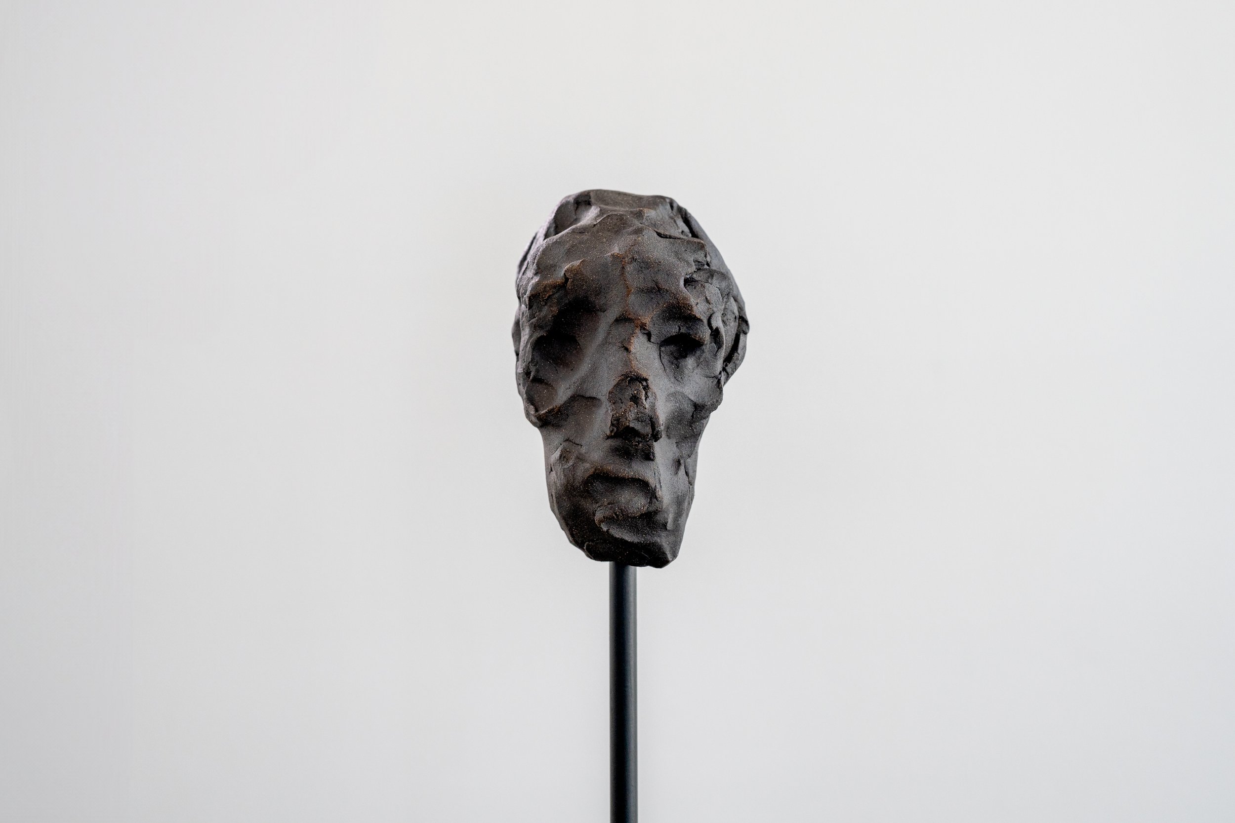 An abstract sculpture of a human face on a black stand, made of dark, textured material, displayed against a plain white background.