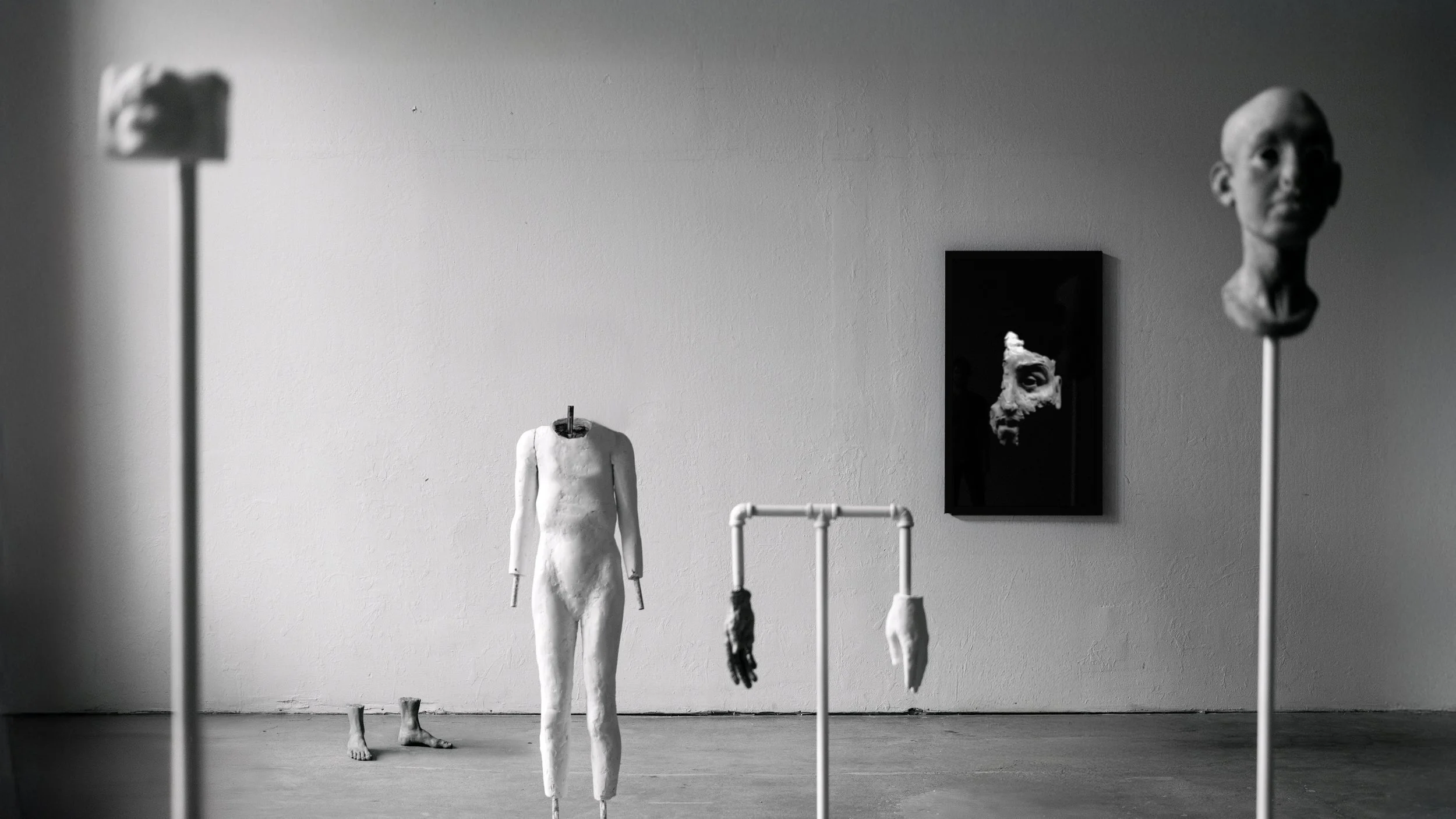 An art exhibit featuring a white mannequin with no arms or face, sculptures of hands, mouth and portrait suspended from pipes, clay feet sit on the ground. On the white wall in the background is a tv screen showing an abstract clay sculpture.