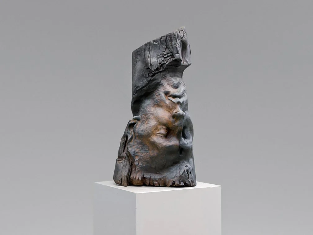 A wooden sculpture with abstract face elements, mounted on a white pedestal against a gray background.
