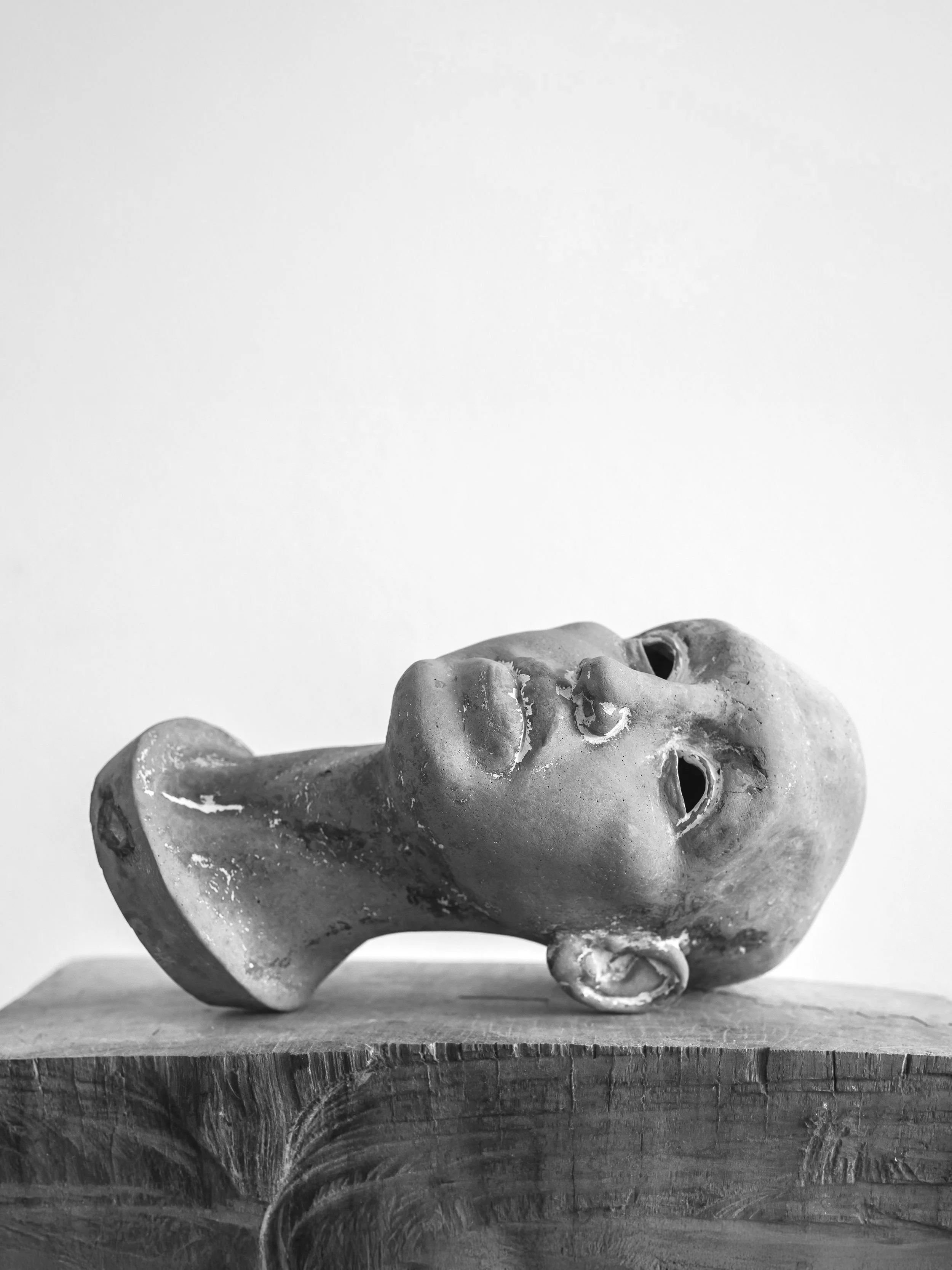 Black and white photograph of a classical sculpture bust of a person's head resting on a wooden surface against a plain background.