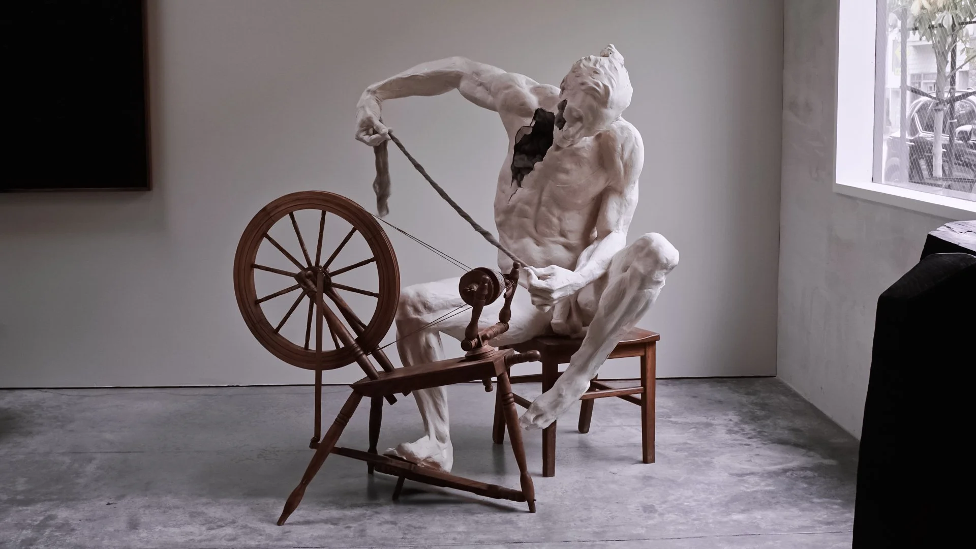 A sculpture of a man with an exaggerated muscular build and long hair, sitting on a chair, pulling a spinning wheel with a string, in a modern art gallery with concrete floor and white walls.