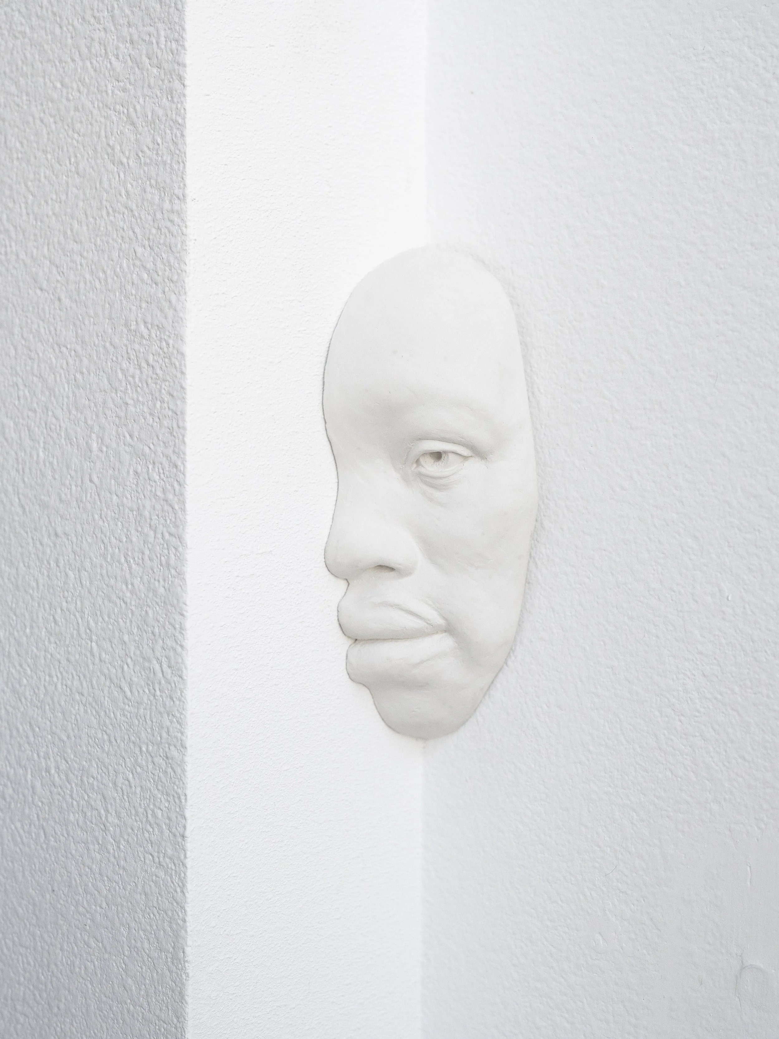 White sculpted face mask emerging from a textured white wall with contrast lighting.
