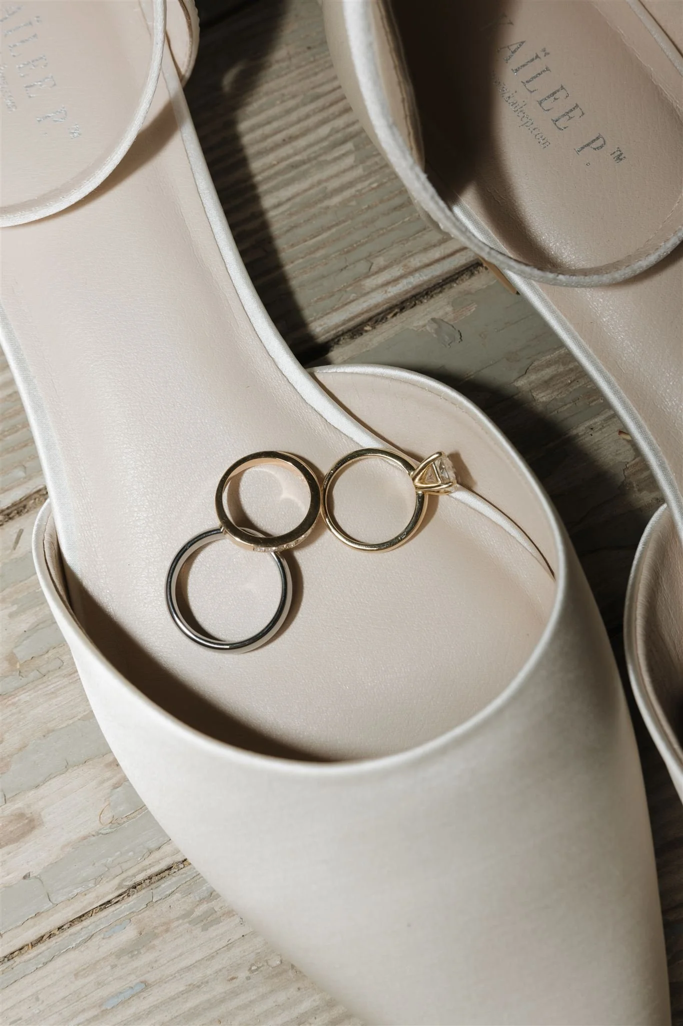 Two wedding rings, one gold with a diamond, placed in an open white shoe box with a pair of white high-heeled shoes with satin finish and stitched edges, on a wooden surface.