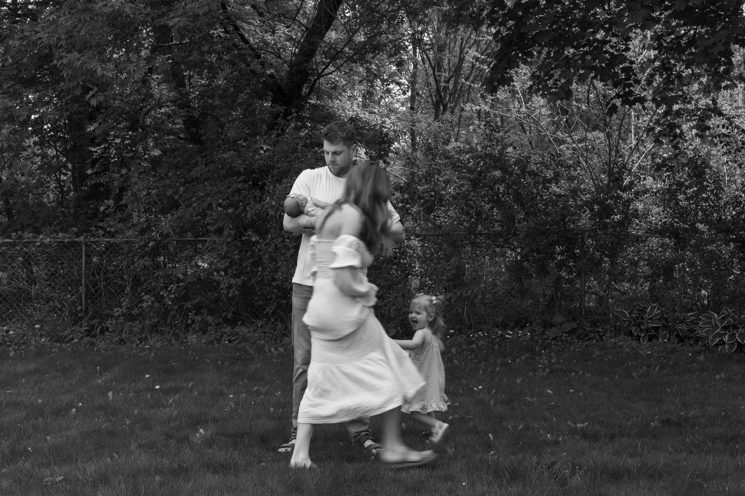 A man and two young girls playing outdoors on a grassy area near trees, with a chain-link fence in the background.