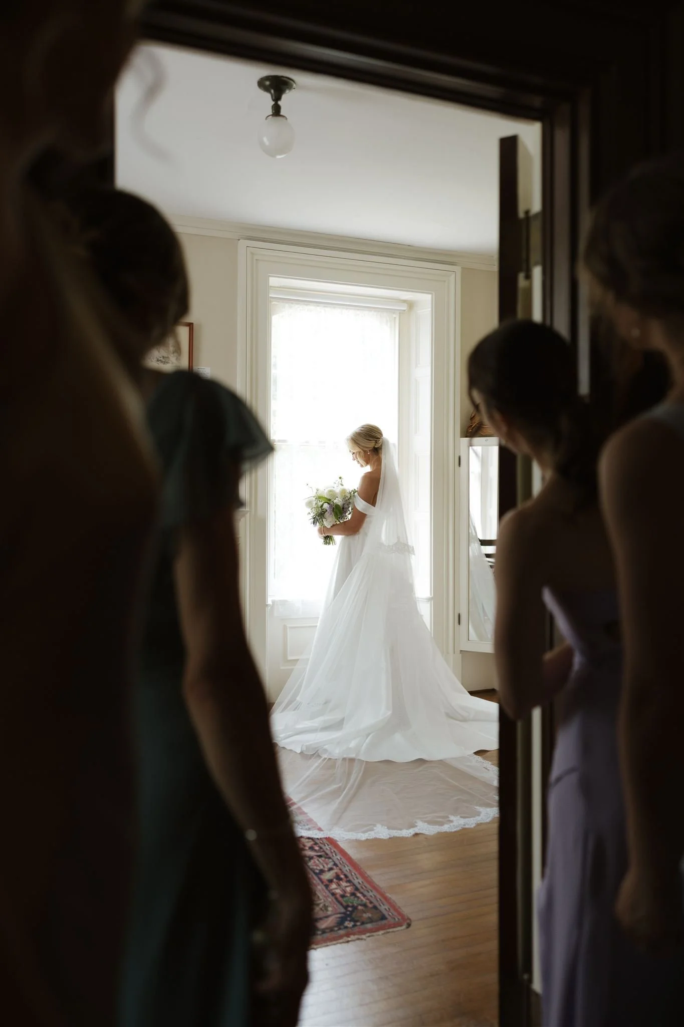 A bride in a white wedding dress and veil holding a bouquet of flowers, standing in front of a large window with natural light, seen through a partially open door with several women watching from the shadows.