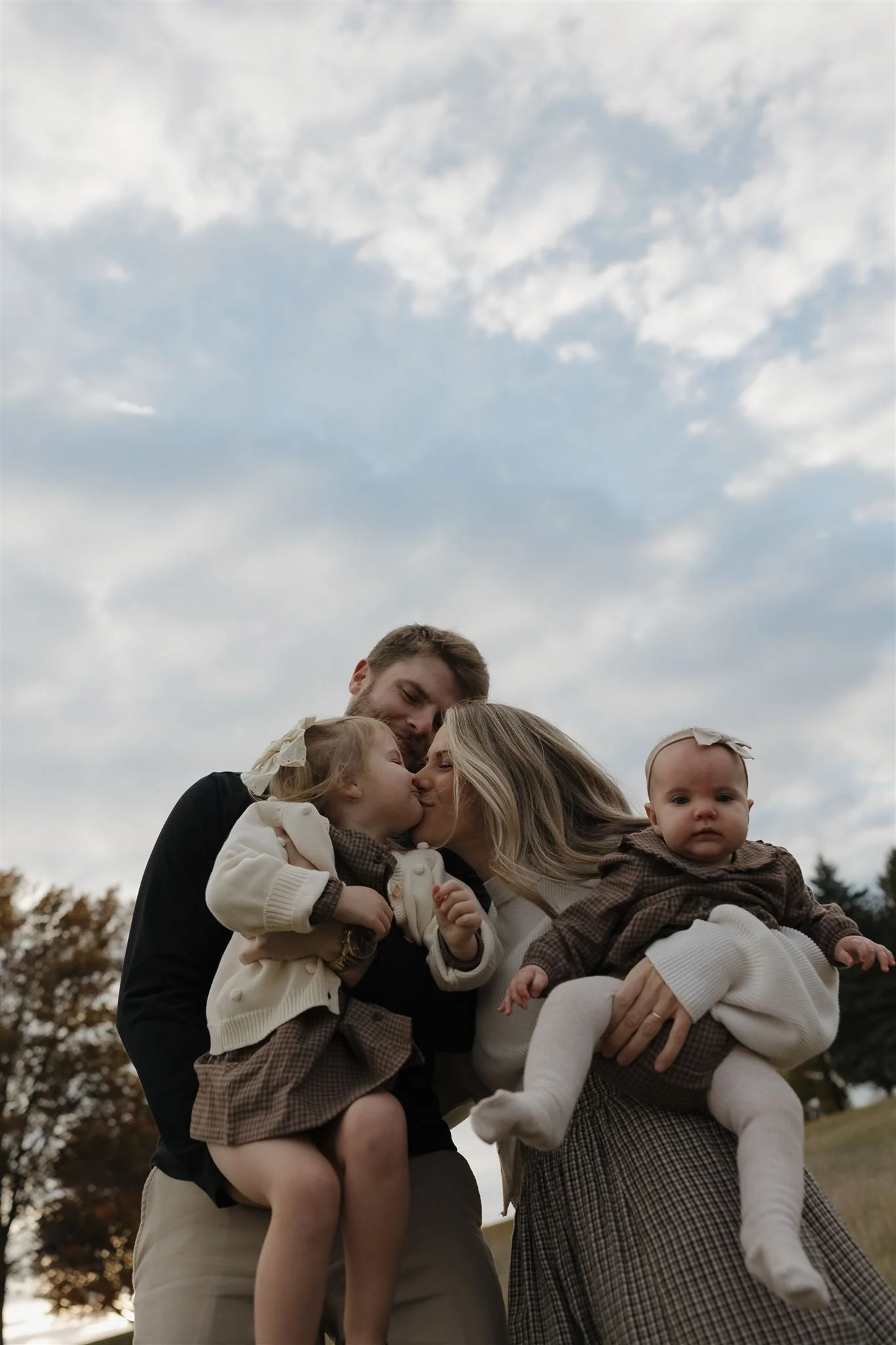 A family of four, a father, mother, and two young girls, sharing a kiss outdoors with a cloudy sky above and trees in the background.