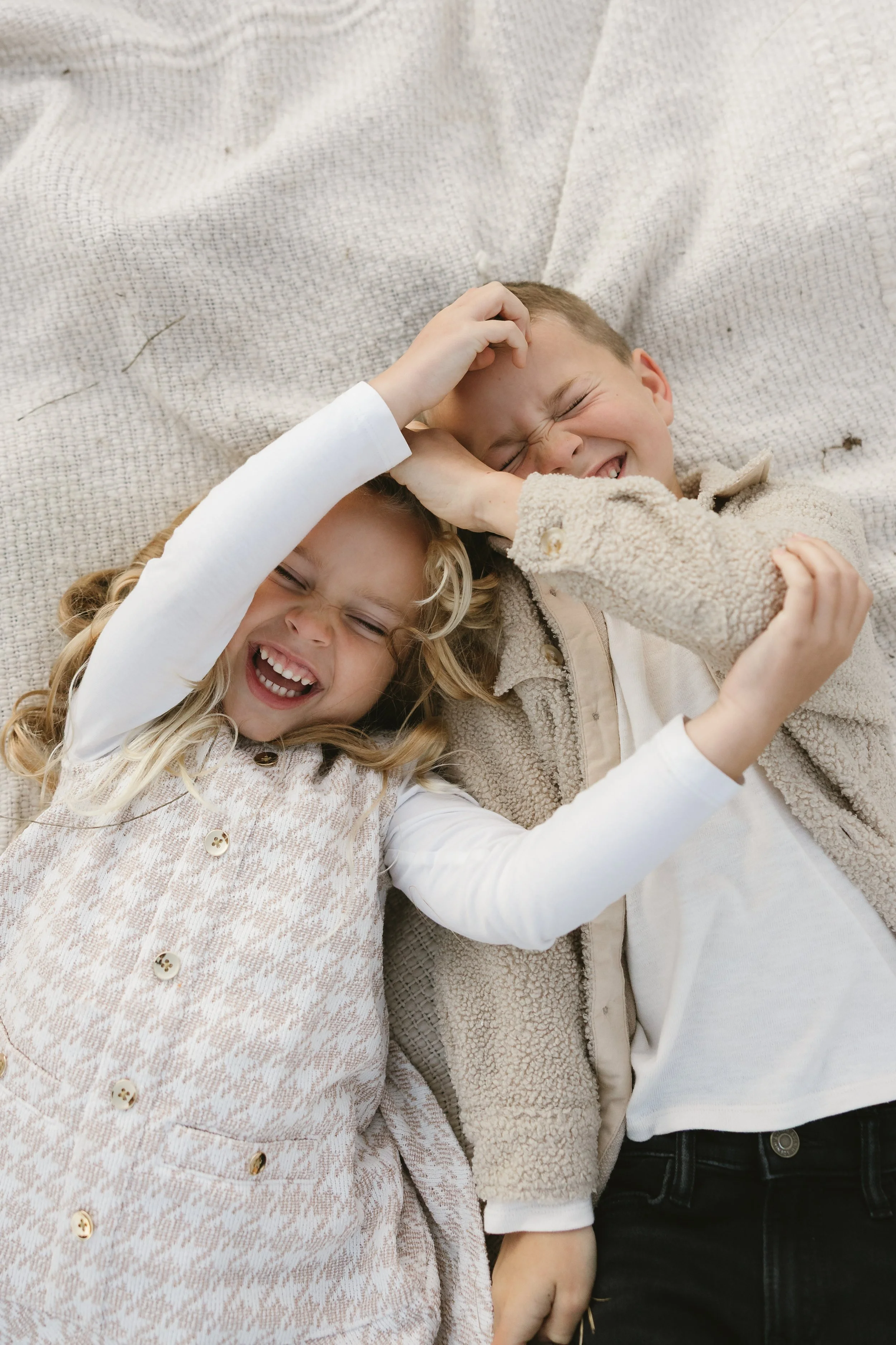Two children, a girl and a boy, are lying on a beige textured blanket, playfully pinching each other's faces and smiling with eyes closed.