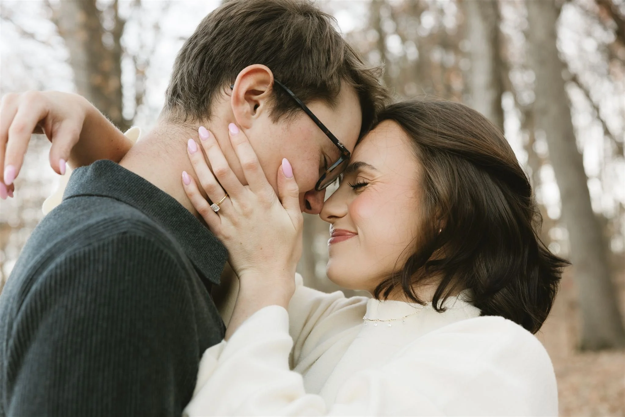 A couple sharing a close, tender moment outdoors in a wooded area, with their foreheads touching and eyes closed, smiling softly.