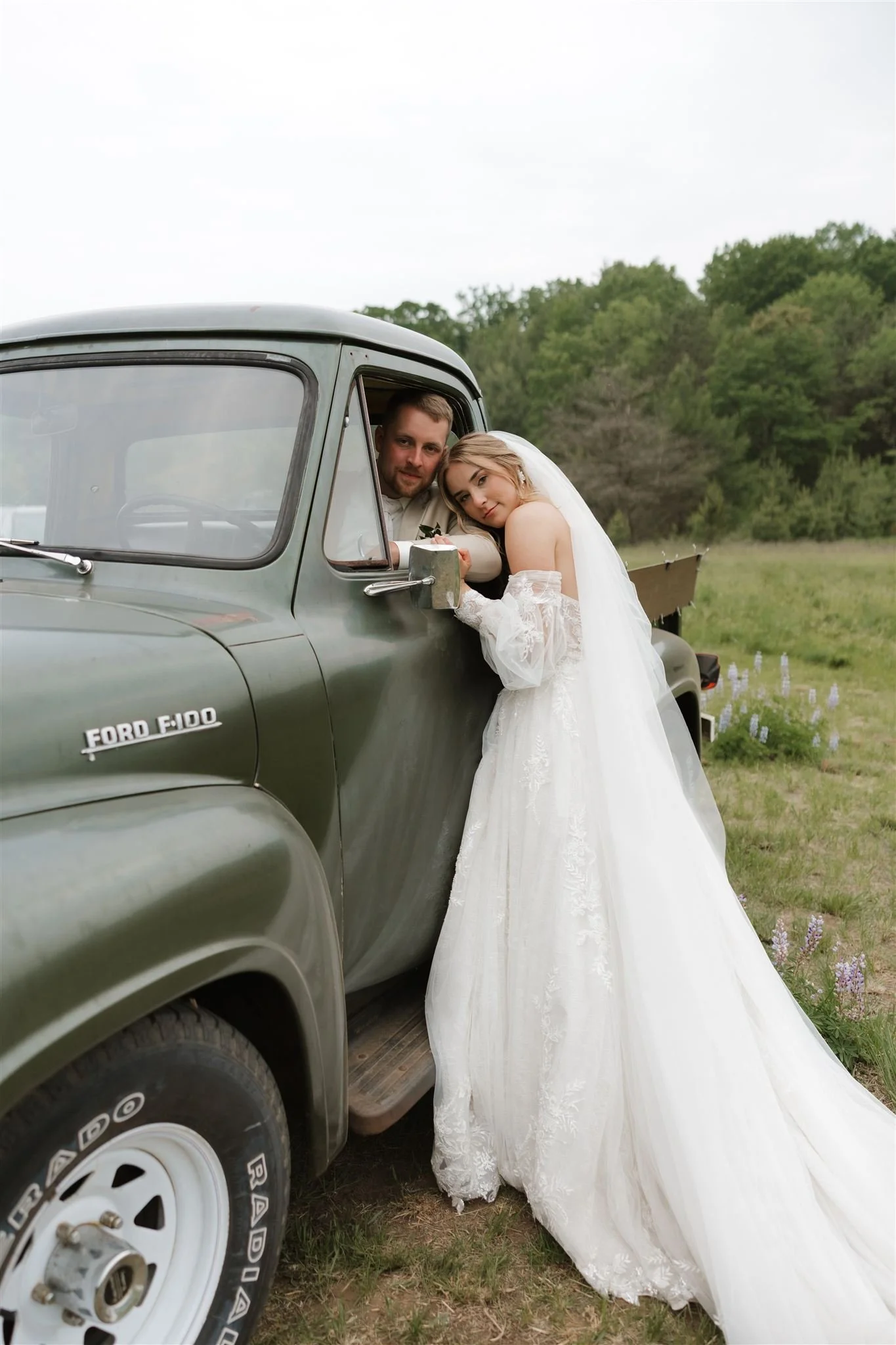 Bride leaning on the side mirror of a vintage green Ford F-100 truck with her arm resting on the window frame, all set in a grassy outdoor field with trees in the background.