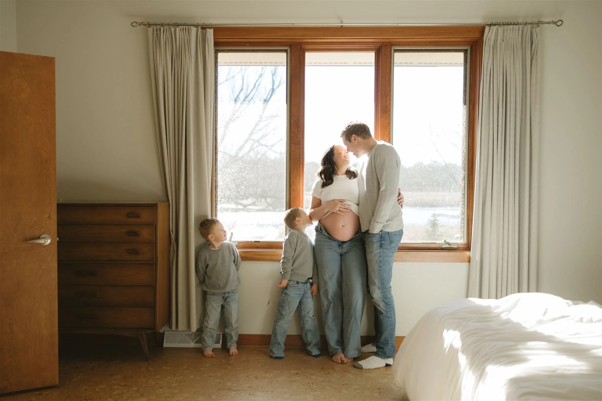A pregnant woman and a man, possibly her partner, standing close together in front of a large window, with two young boys standing on either side of them. The woman is touching her belly and smiling at the man. The scene is bright and warm, with a snowy landscape visible outside.