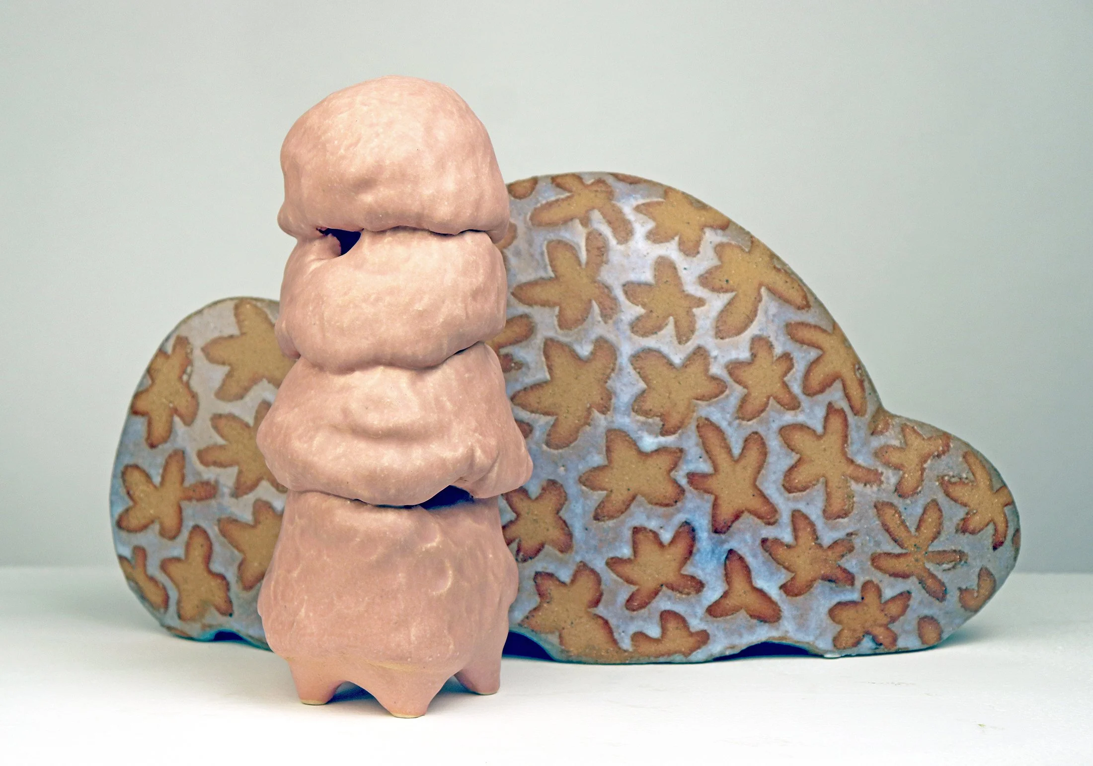 A fleshy pink ceramic form sits in from of an abstract shape glazed with a pattern of flowers. It is an abstract sculpture that recalls a human body in front of a sofa or other soft furnishing.