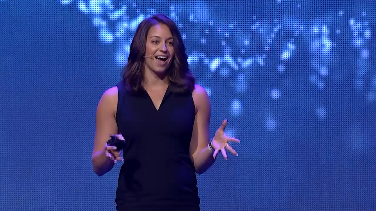 Lo Myrick, keynote speaker. A woman in a sleeveless black dress giving a presentation on a stage with a blue and white sparkly background.