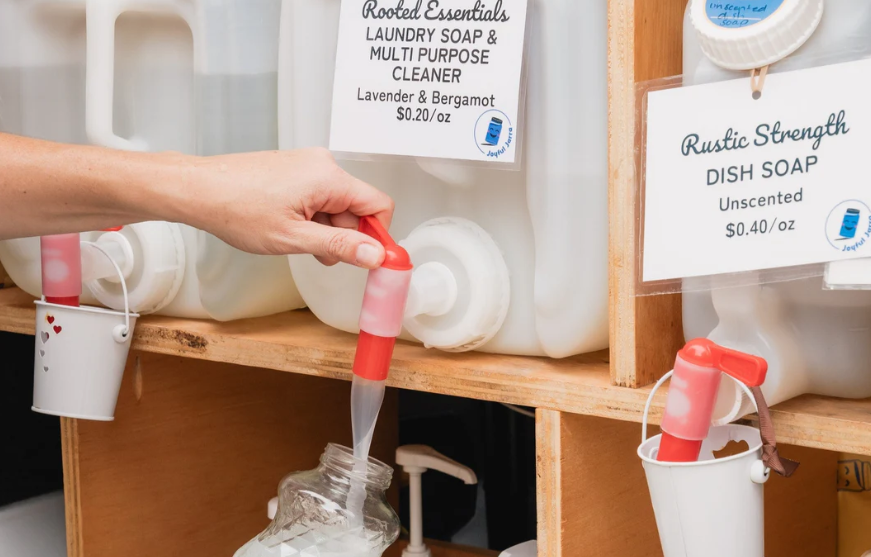 Close-up photo of someone refilling a container with laundry detergent. There are several large refillable containers of soap products on the shelf. Source: Joyful Jarra.