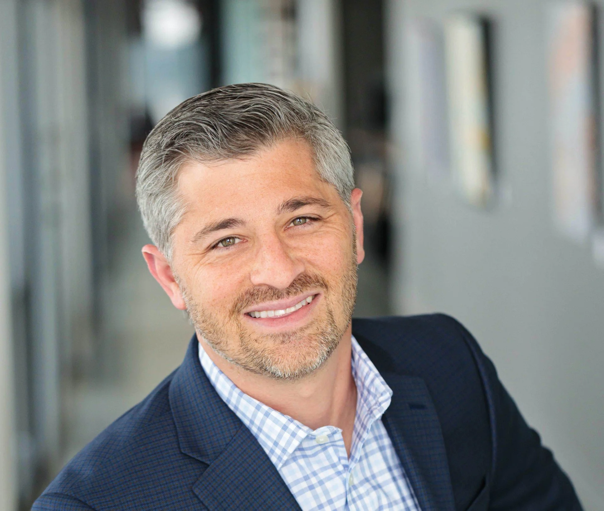 Headshot of Adrian Sasine, the author of the article. Adrian is smiling into the camera and wearing a blue plaid blazer and a blue and white plaid shirt. The background is out of focus and gray. Photo Credit: Adrian Sasine.