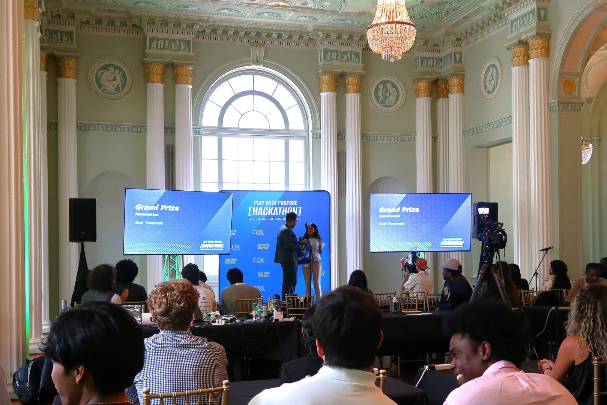 Photo from the Play with Purpose Sustainability Hackathon award ceremony where two people are on stage in front of blue screens and an audience of students faces the stage and winners..  Source: Pearl Kaplan/Tech Square ATL.