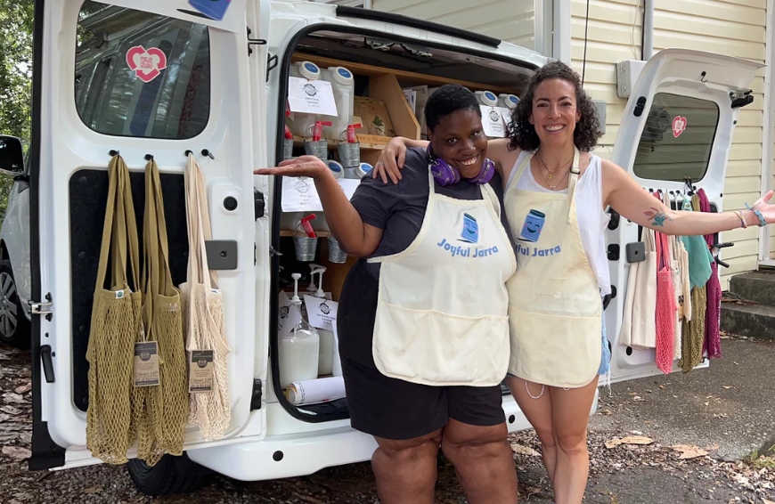 Photo of Veronica Apecena and a Joyful Jarra employee smiling and standing in front of the mobile refillery. There are various bags, refillable containers in the photo. Source: Joyful Jarra.