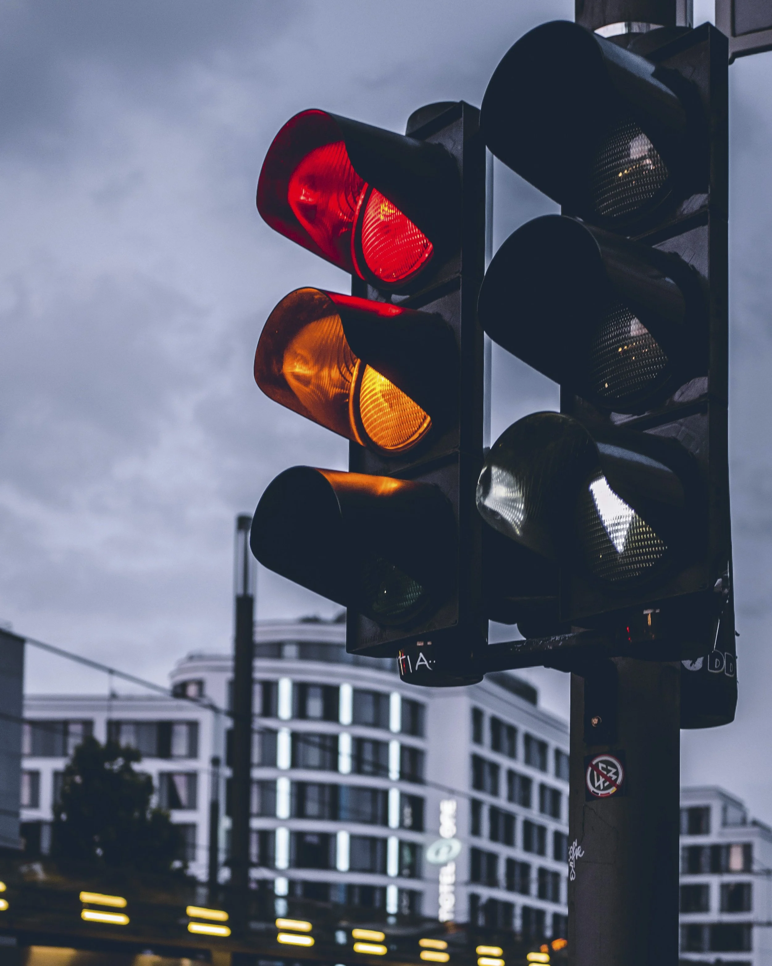 Stock photo of a traffic signal on a city street with mid-rise modern buildings in the background. The sky is gray and the signal is showing orange and red lights. Source: Maxim Abramov/Unsplash.