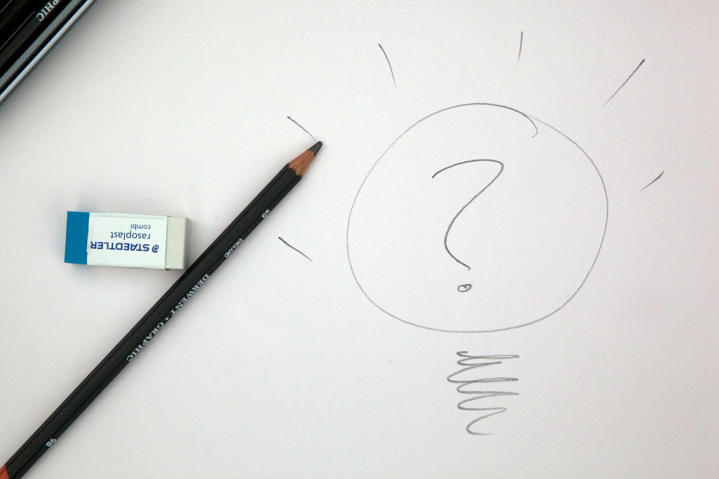 Image shows a charcoal pencil and eraser on a white piece of drawing paper. There is a lightbulb with a question marked sketched on the paper. Photo Credit: Mark Fletcher Brown via Unsplash.