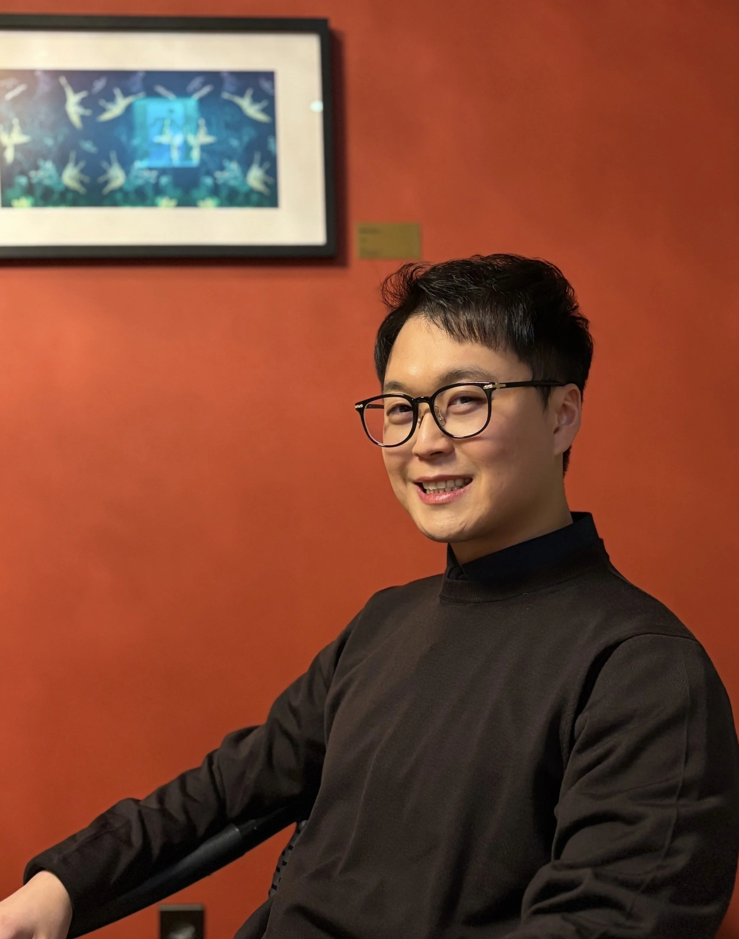 Portrait style photo of Seung Jae Lieu sitting in front of a orange/burnt red background. Photo: Seung Jae Lieu.