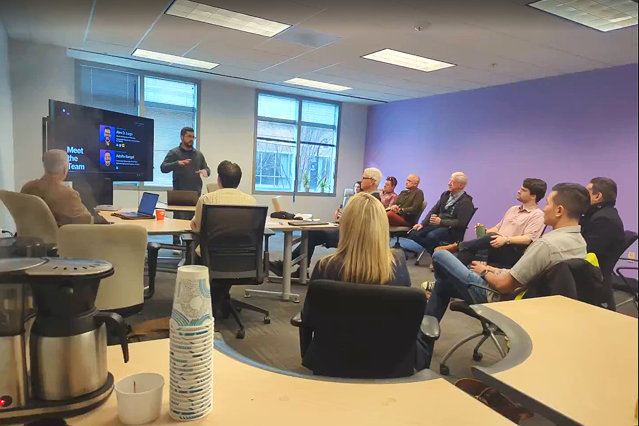 Image shows a room full of people watching a presentation at ATDC's Mond(AI)y Coffee. The presenter is standing to the right of a TV screen. A coffee pot and cups are in the bottom left corner on a desk. Credit: Mond(AI)y Coffee.