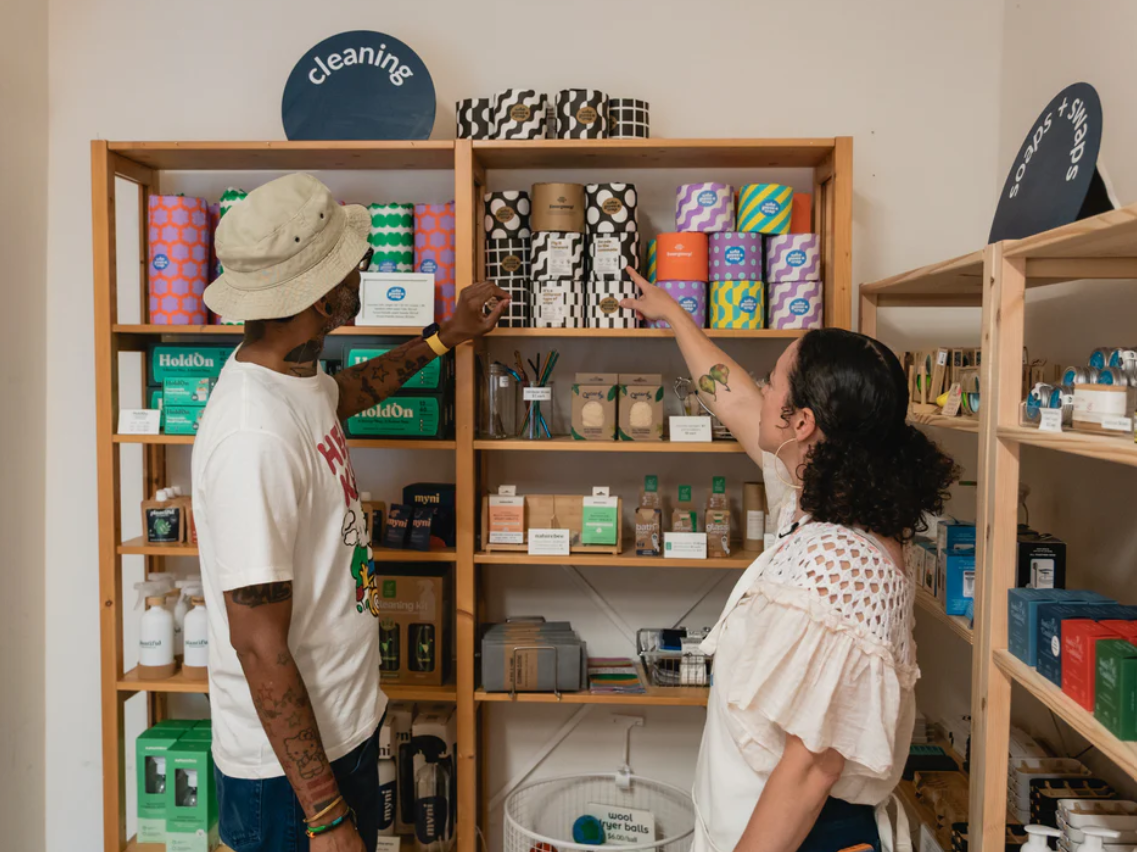 Photo of Veronica Apecena showing a Joyful Jarra visitor a shelf full of non-toxic and low-waste cleaning products. There are bar soaps without plastic wrappers, for example. Source: Joyful Jarra.
