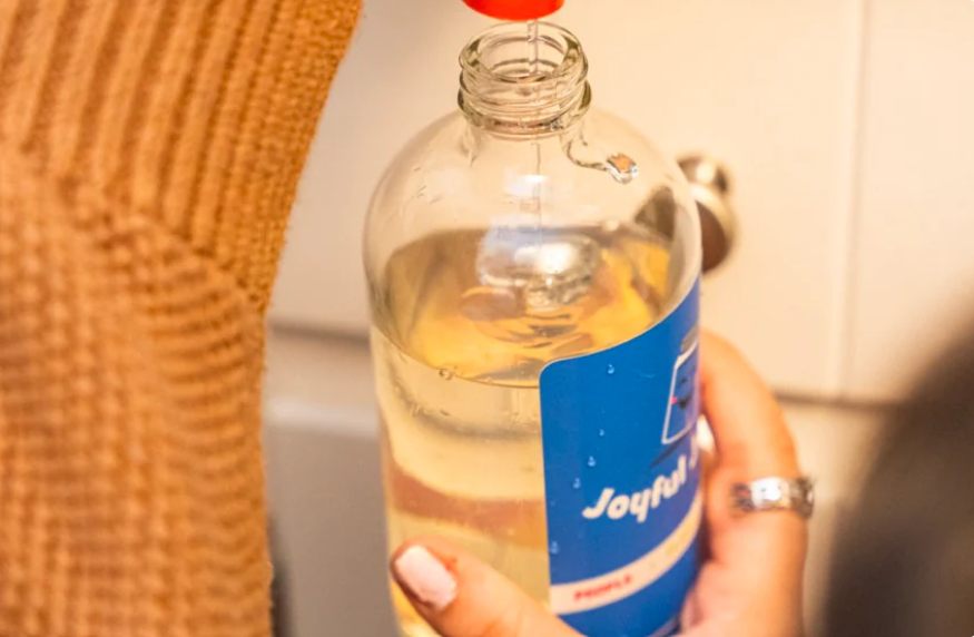 Close-up photo of someone refilling a smaller glass container with soap. The person holding the glass container is wearing an orange sweater. Source: Joyful Jarra.