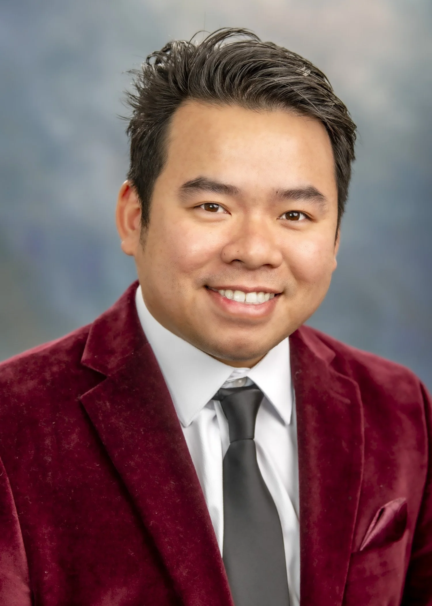 Portrait photo of Dr. Ethan Trinh, CEO and Founder of Dr. Trinh Foundation. Dr. Trinh is wearing a dark red blazer, white shirt, and a gray tie. The background is a light blue-gray. Photo Credit: Dr. Trinh Foundation.