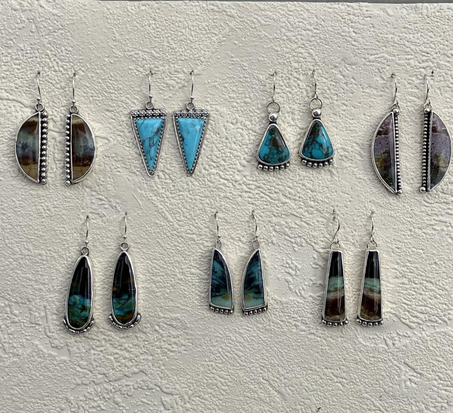 Collection of ten pairs of silver earrings with various turquoise and multicolored stone inlays hanging on a textured beige background.