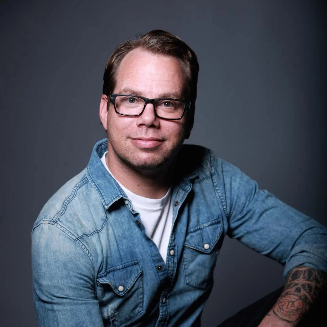 A young man with glasses, wearing a denim shirt and a white t-shirt, smiling and seated against a plain dark background.