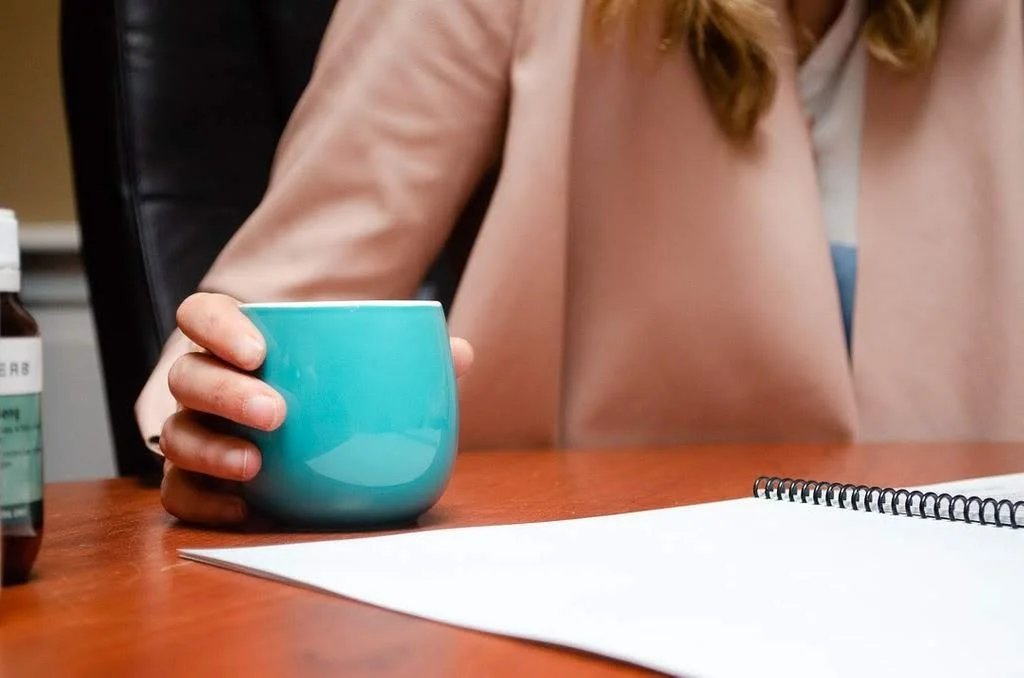 Dr. Katie ND sitting at a desk holding a blue mug in their right hand. There is a bottle of supplement on the left and a spiral notebook on the right on the wooden desk.